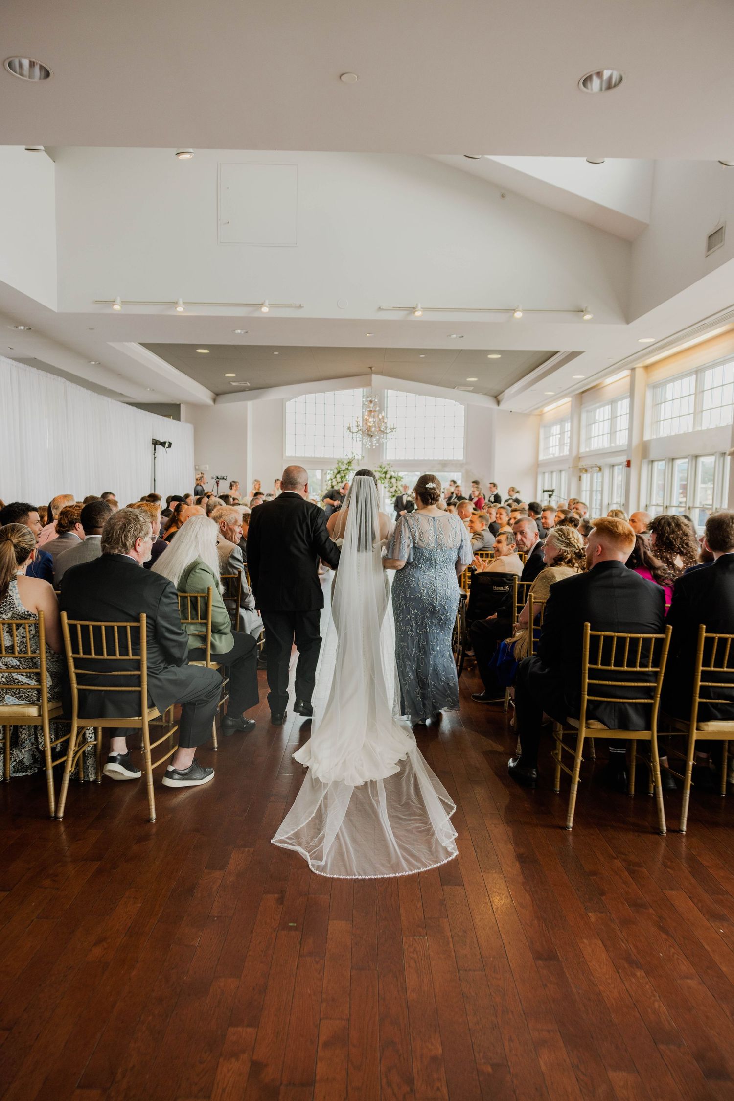 Bride walking down aisle in elegant gown with long veil at indoor wedding ceremony.
