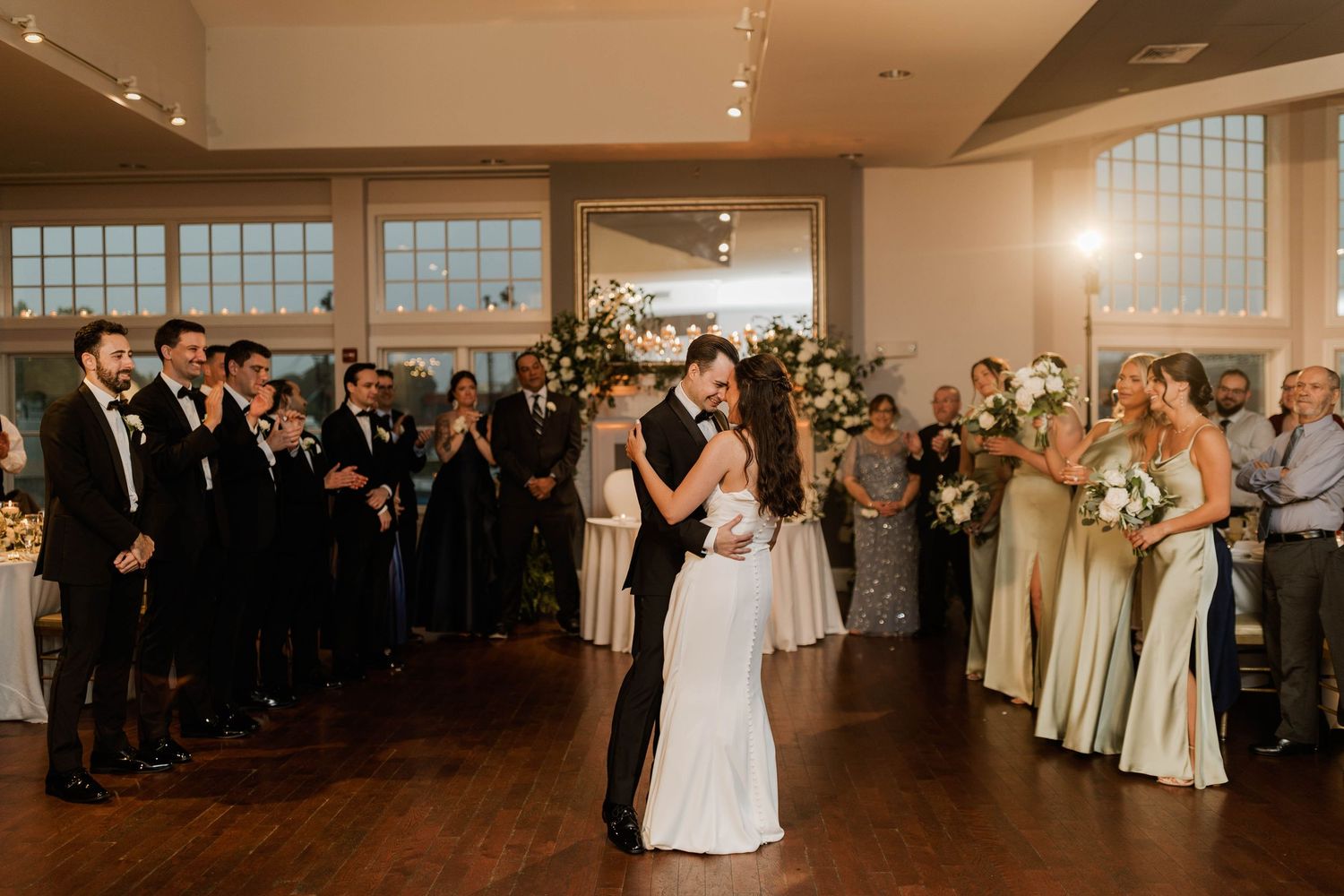 Bride and groom sharing first dance surrounded by applauding guests in elegant wedding reception venue.