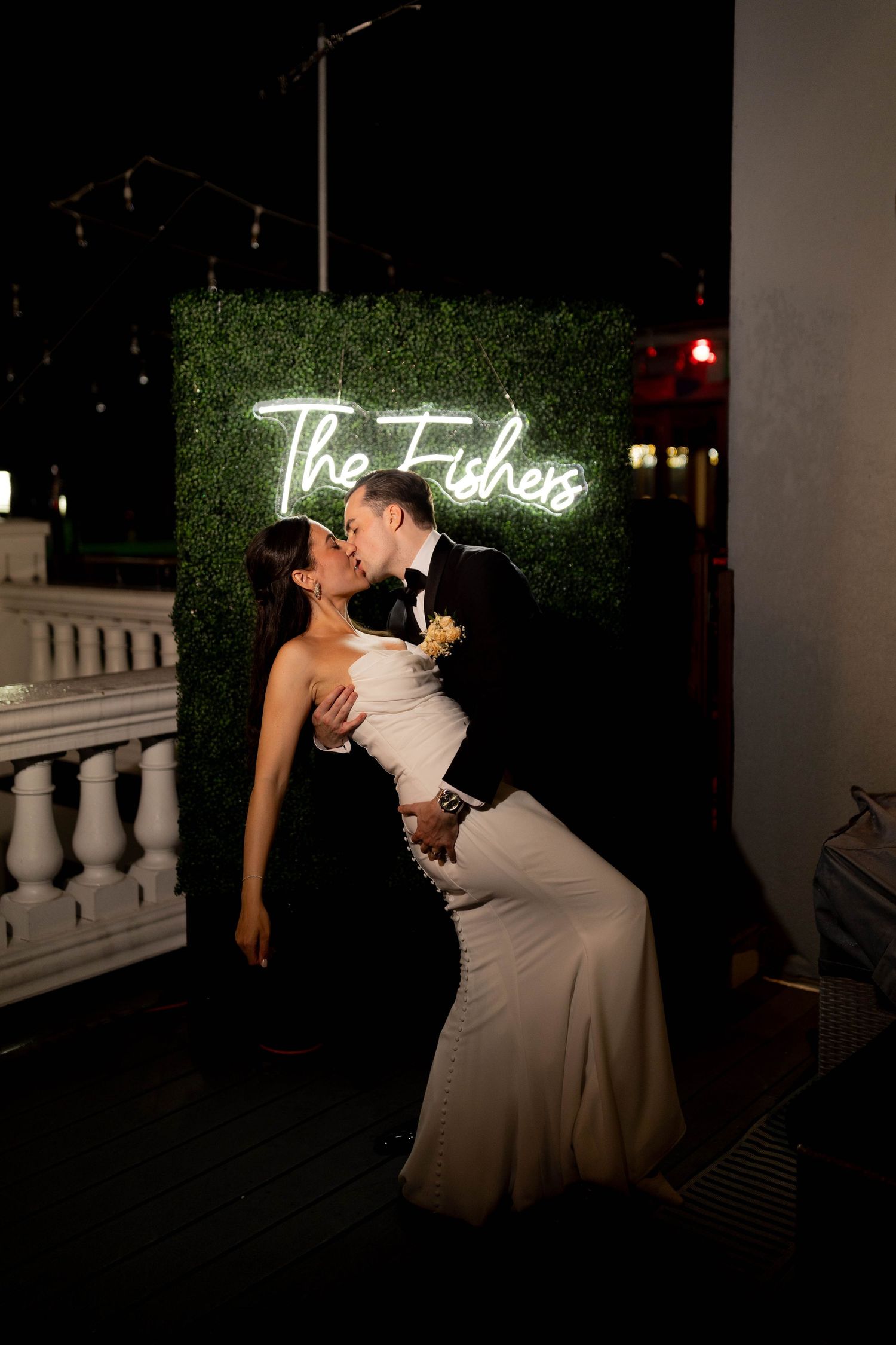 Bride and groom kiss romantically in front of illuminated The Fishers neon sign at night.
