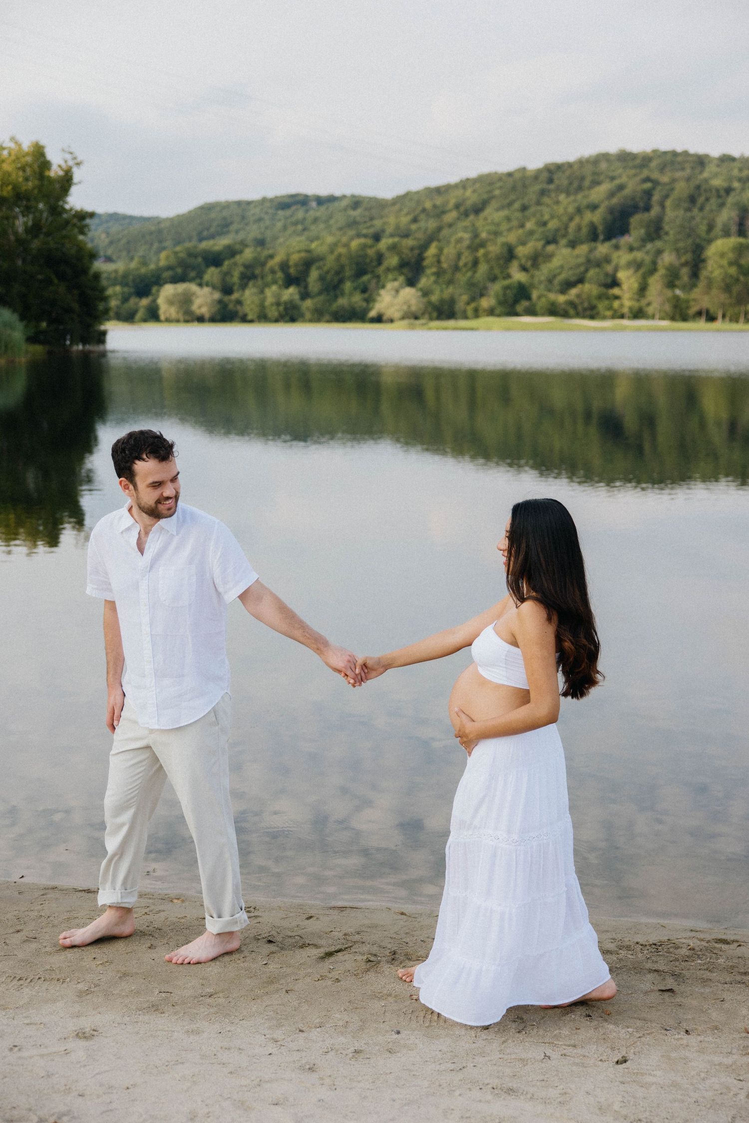 Pregnant couple in white outfits holding hands by a serene lake with lush green hills in the background.