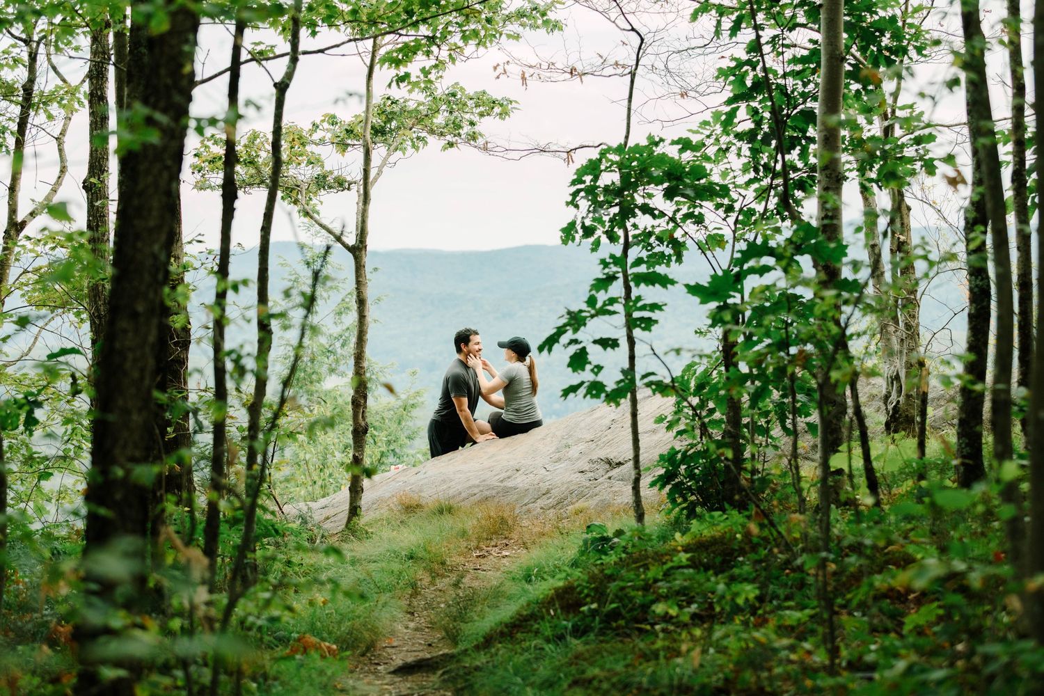 Couple sitting on a rock ledge surrounded by lush green forest trees with misty mountain views in background.