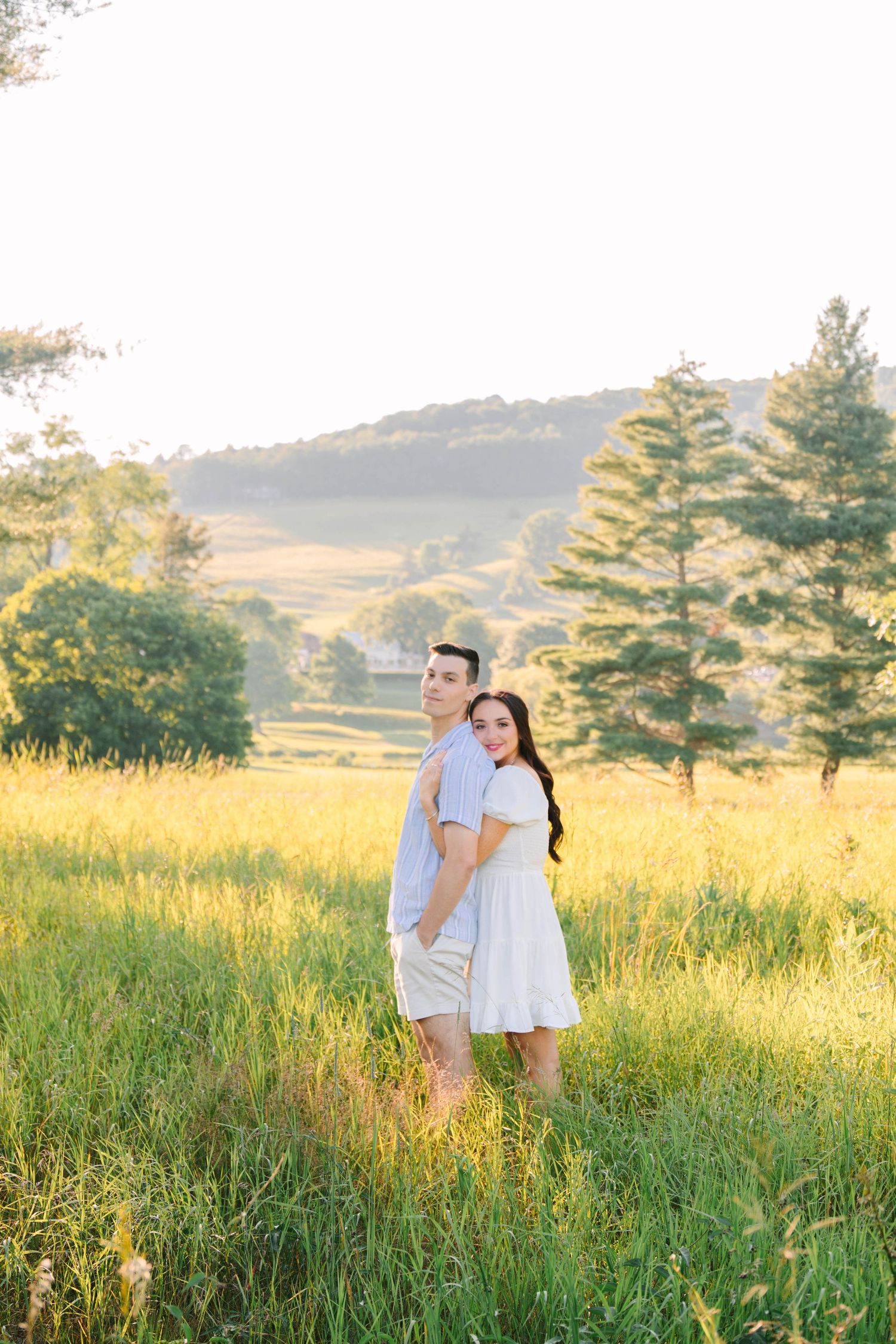 Couple standing in golden sunlit field, woman in white dress embracing man from behind with rolling hills backdrop.