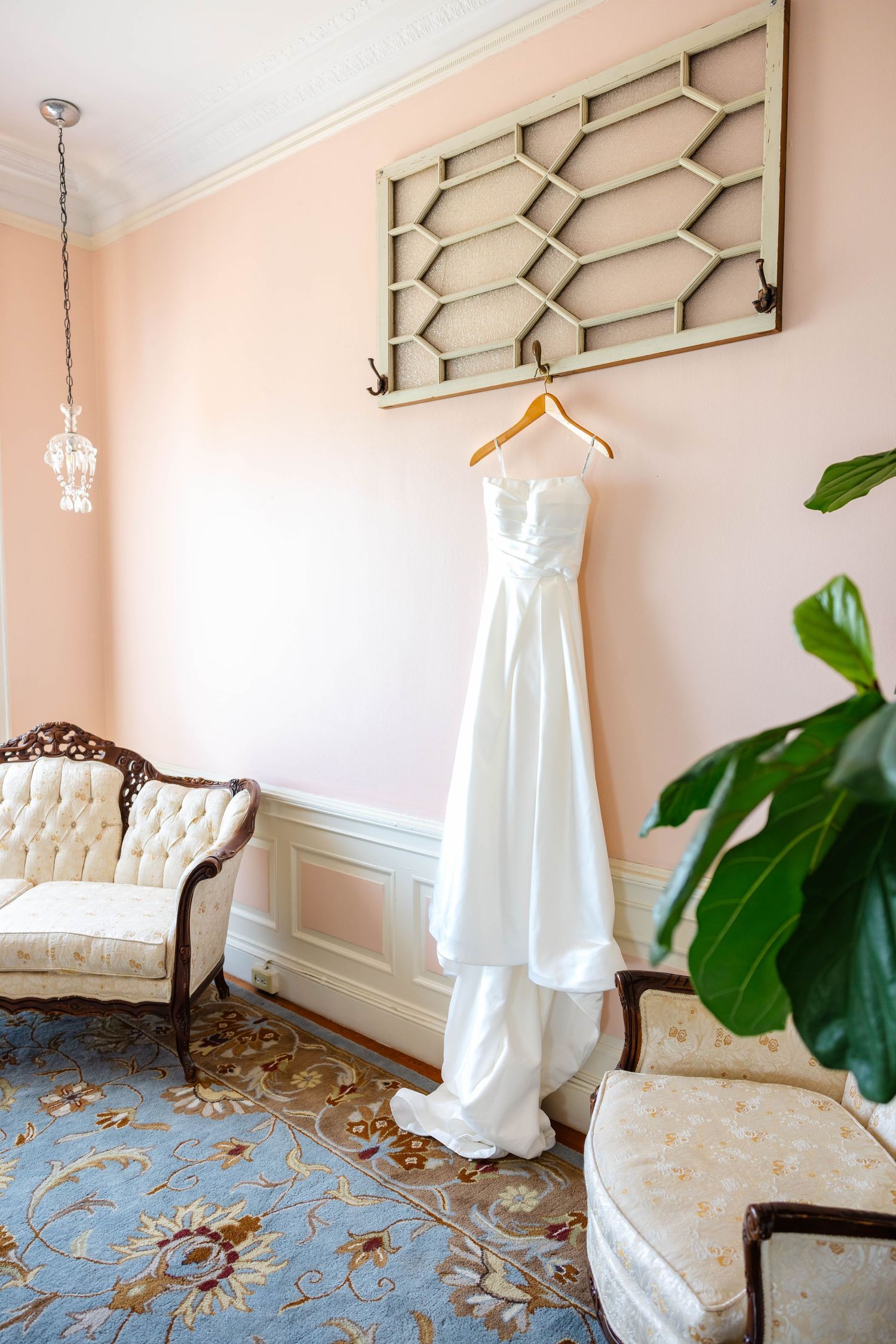 White wedding dress hanging on wooden hanger against pink wall in elegant vintage room with tufted sofa.