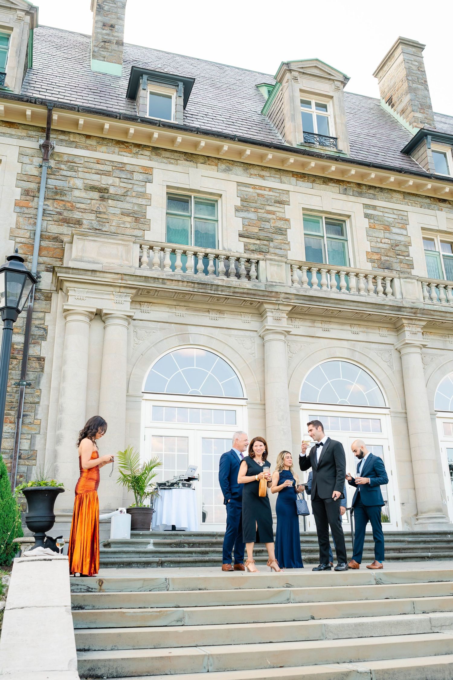 Wedding guests socializing on steps outside a grand stone mansion venue.