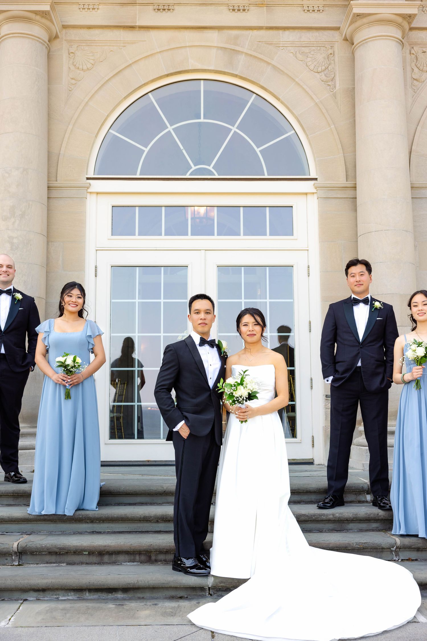 Wedding party posing on steps of elegant neoclassical building, bride in white gown with blue bridesmaids.