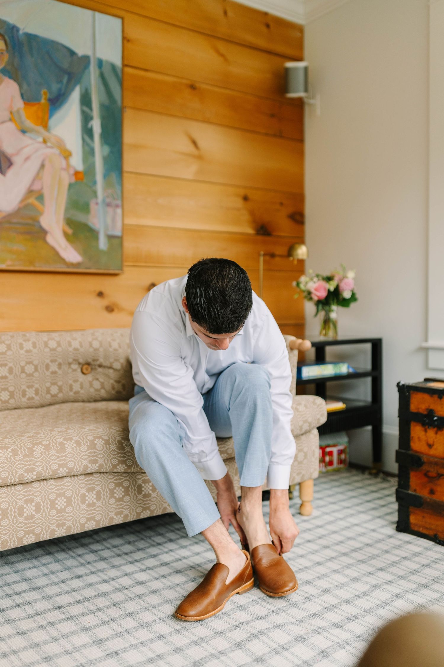 Groom in white shirt and light blue pants putting on brown leather loafers while seated on a patterned sofa.