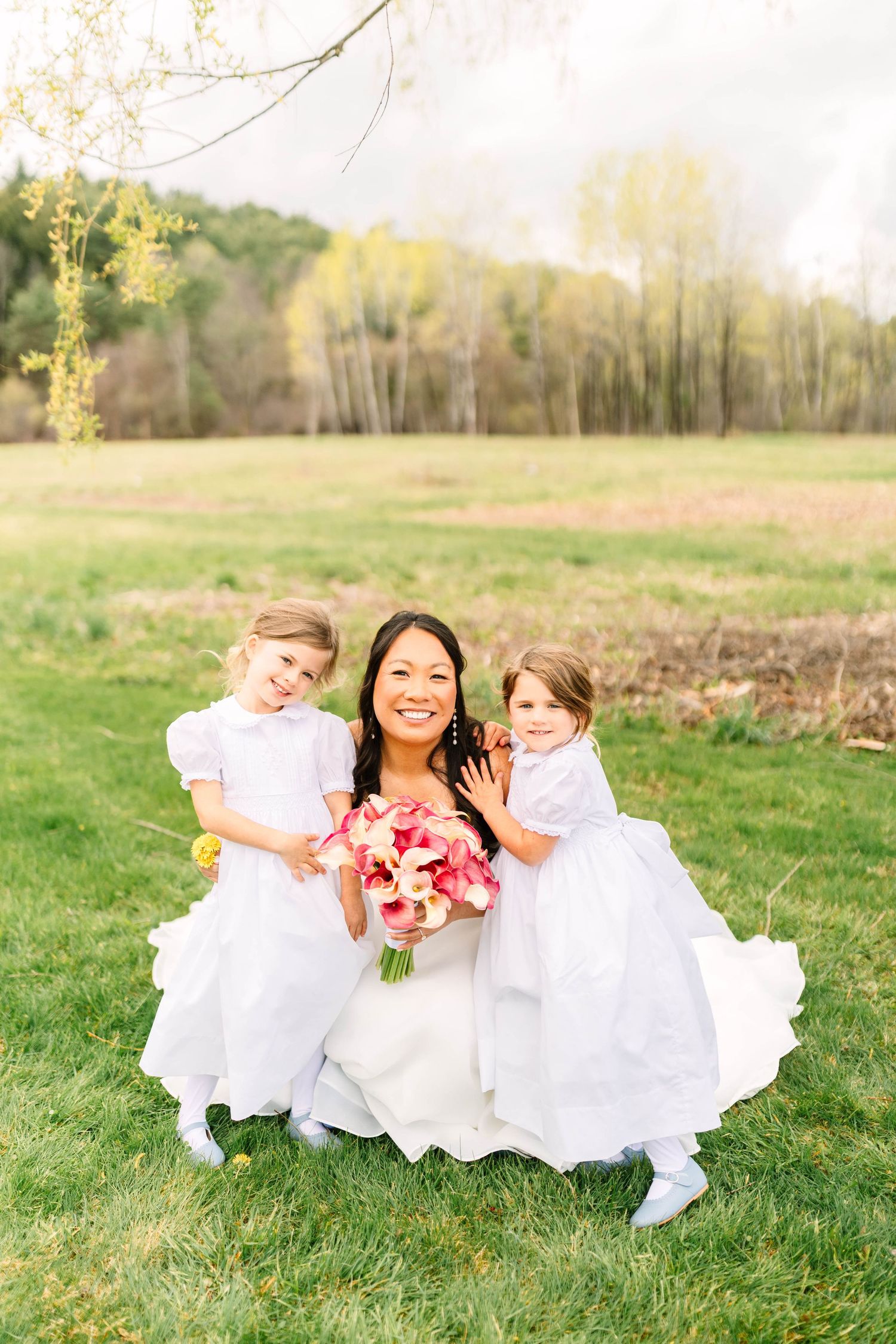 Smiling bride in white gown crouching with two flower girls holding a pink bouquet in a green field.