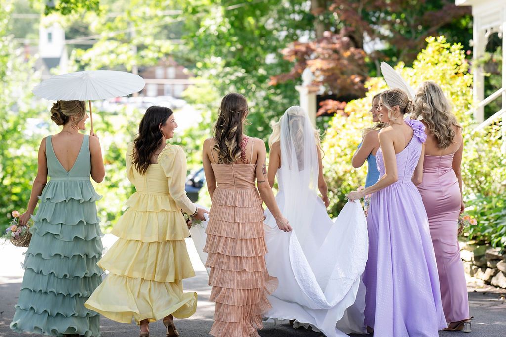 Five bridesmaids in colorful pastel gowns walk with a bride in a white wedding dress and veil outdoors.