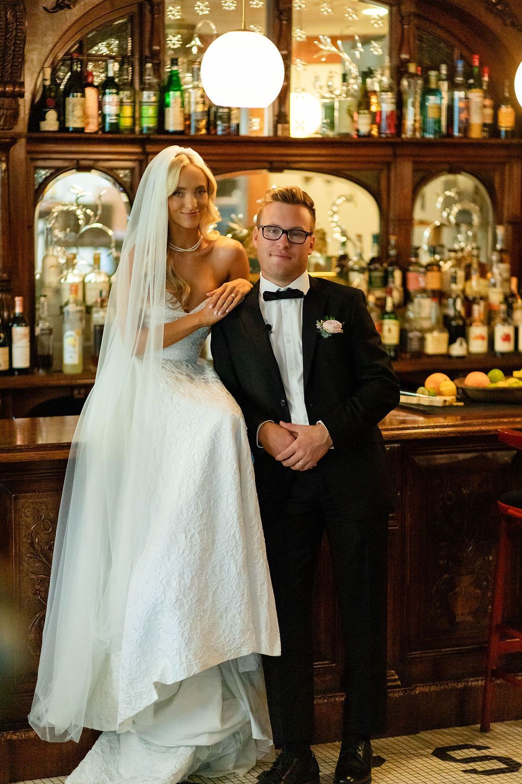 Bride and groom posing together at an elegant vintage wooden bar on their wedding day.