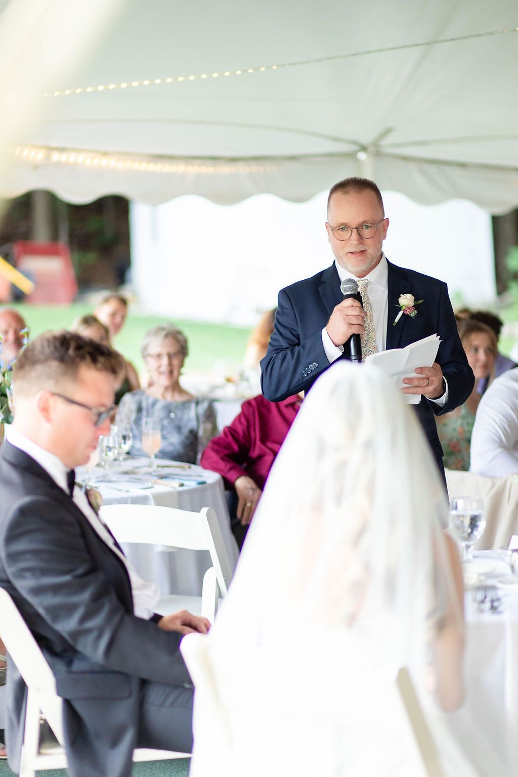 Father of bride delivers heartfelt speech with microphone at elegant tented wedding reception.