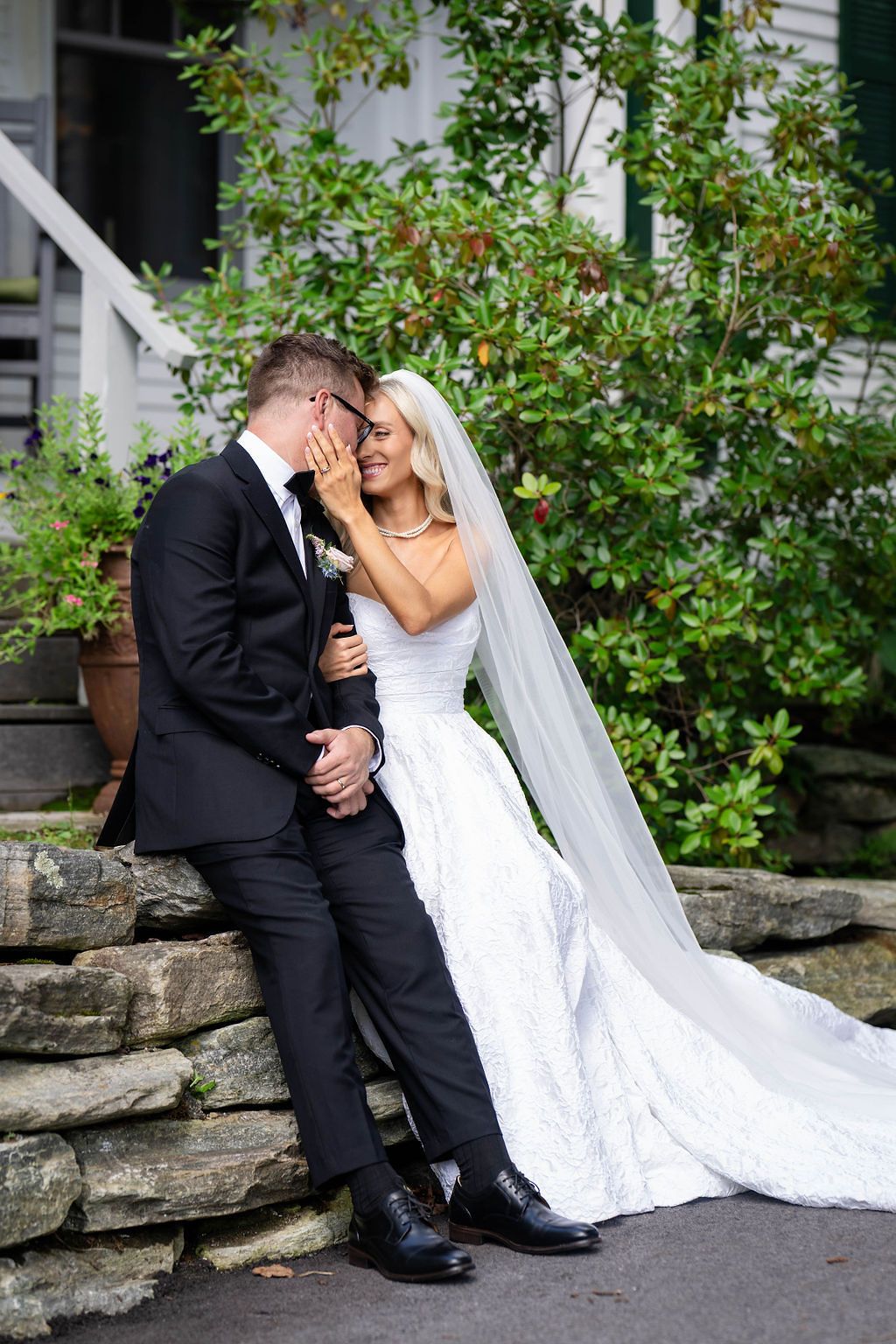 Bride and groom share an intimate moment seated on a stone wall outside a white house surrounded by lush greenery.