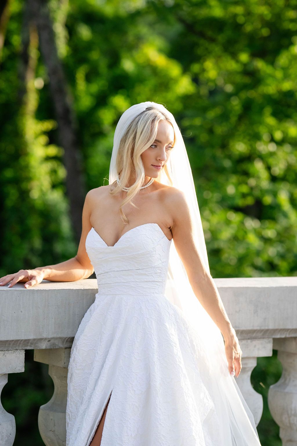 Elegant blonde bride in strapless white gown and veil posing on a stone balcony amid greenery.