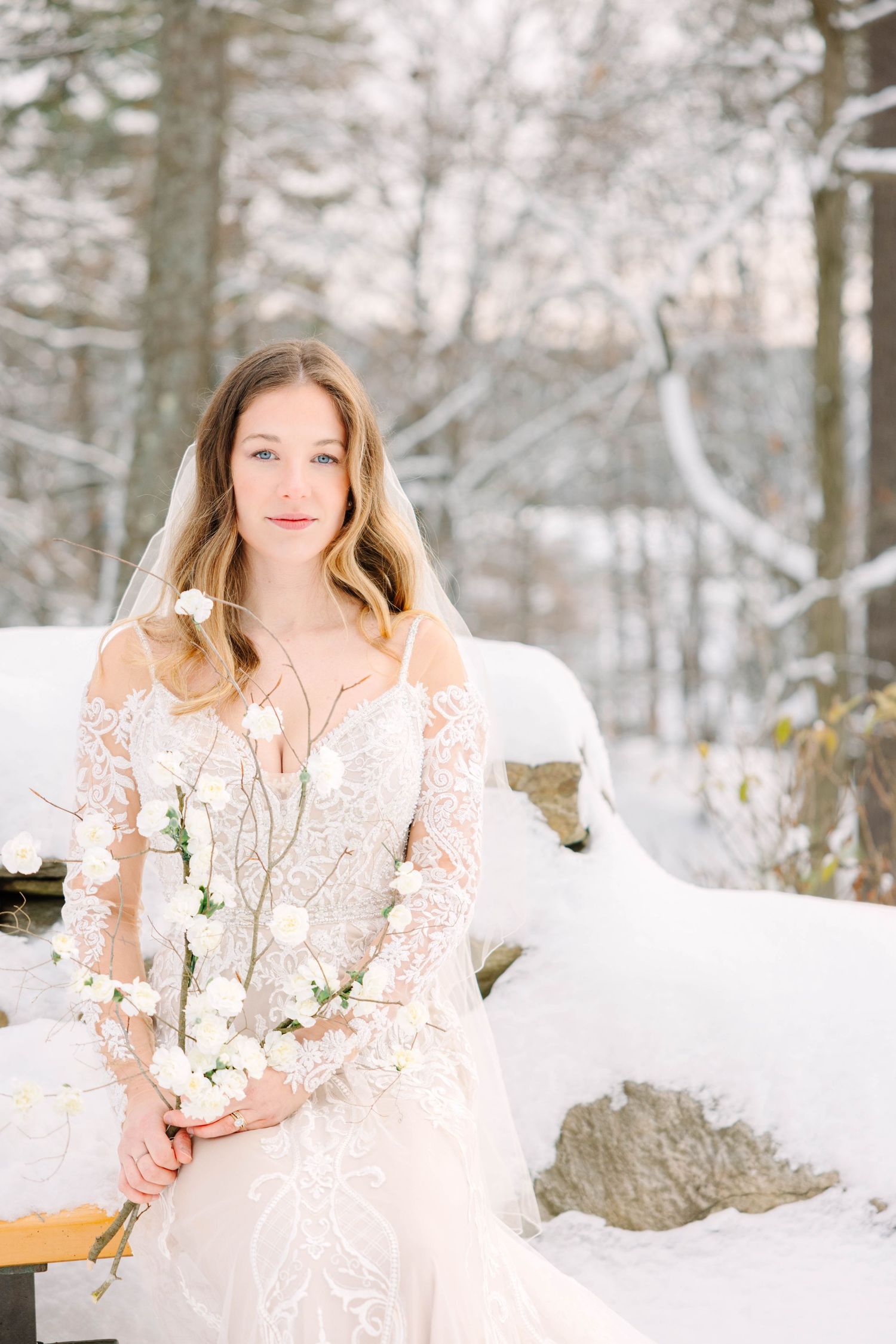 Bride in lace wedding dress and veil sits outdoors in snowy winter setting holding white flowers.