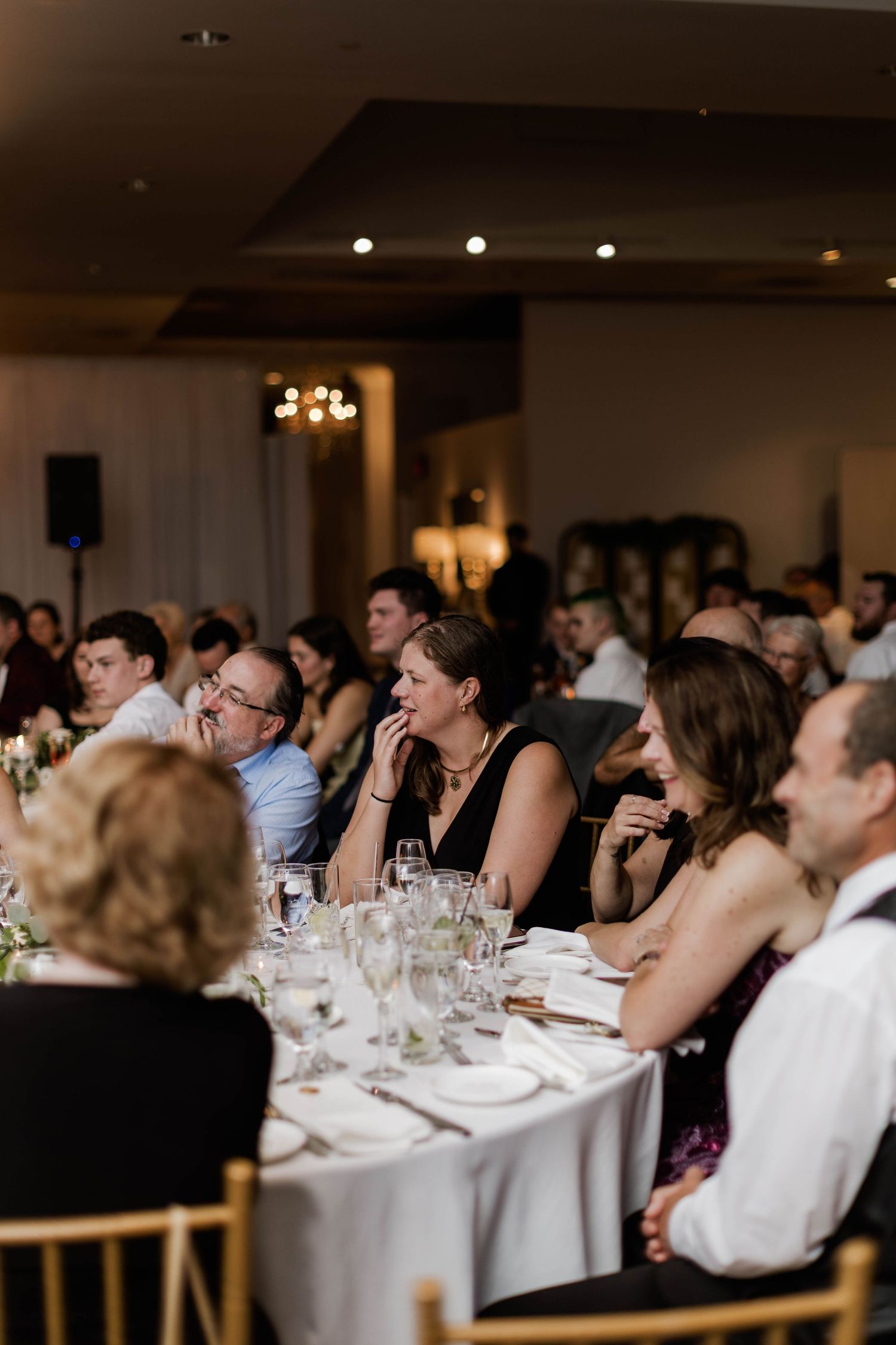 Wedding reception guests attentively listening to speeches at elegantly set tables in a warmly lit venue.