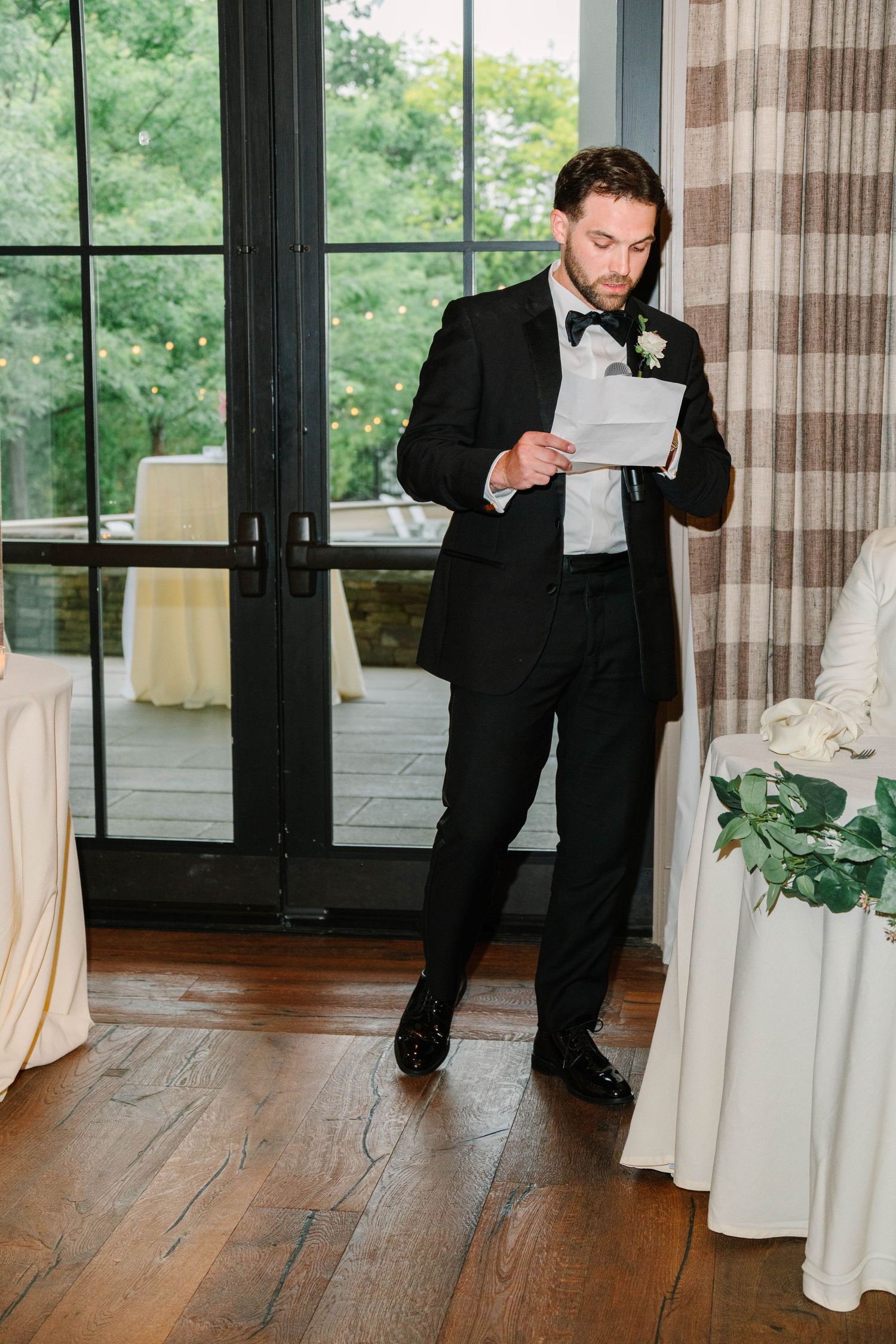 Groom in black tuxedo reads handwritten notes during wedding reception speech indoors.