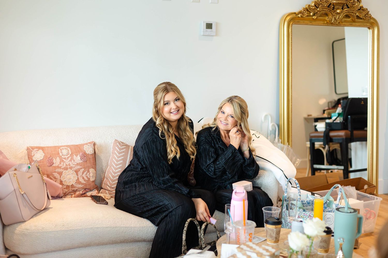 Two bridesmaids in black robes smiling on pink couch with gold mirror and beauty products nearby.
