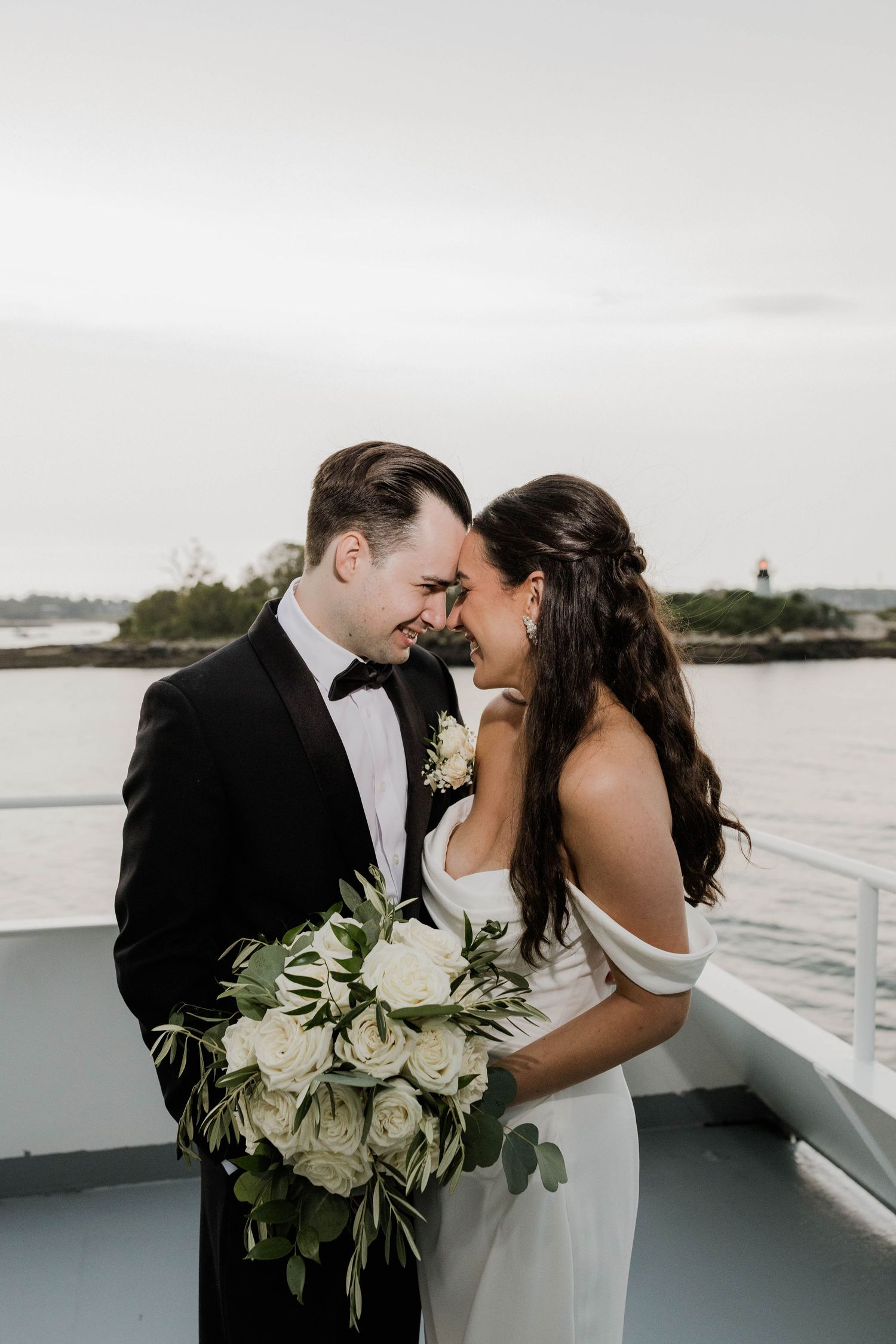 Bride and groom share intimate moment on boat deck, groom holding white rose and greenery bouquet.