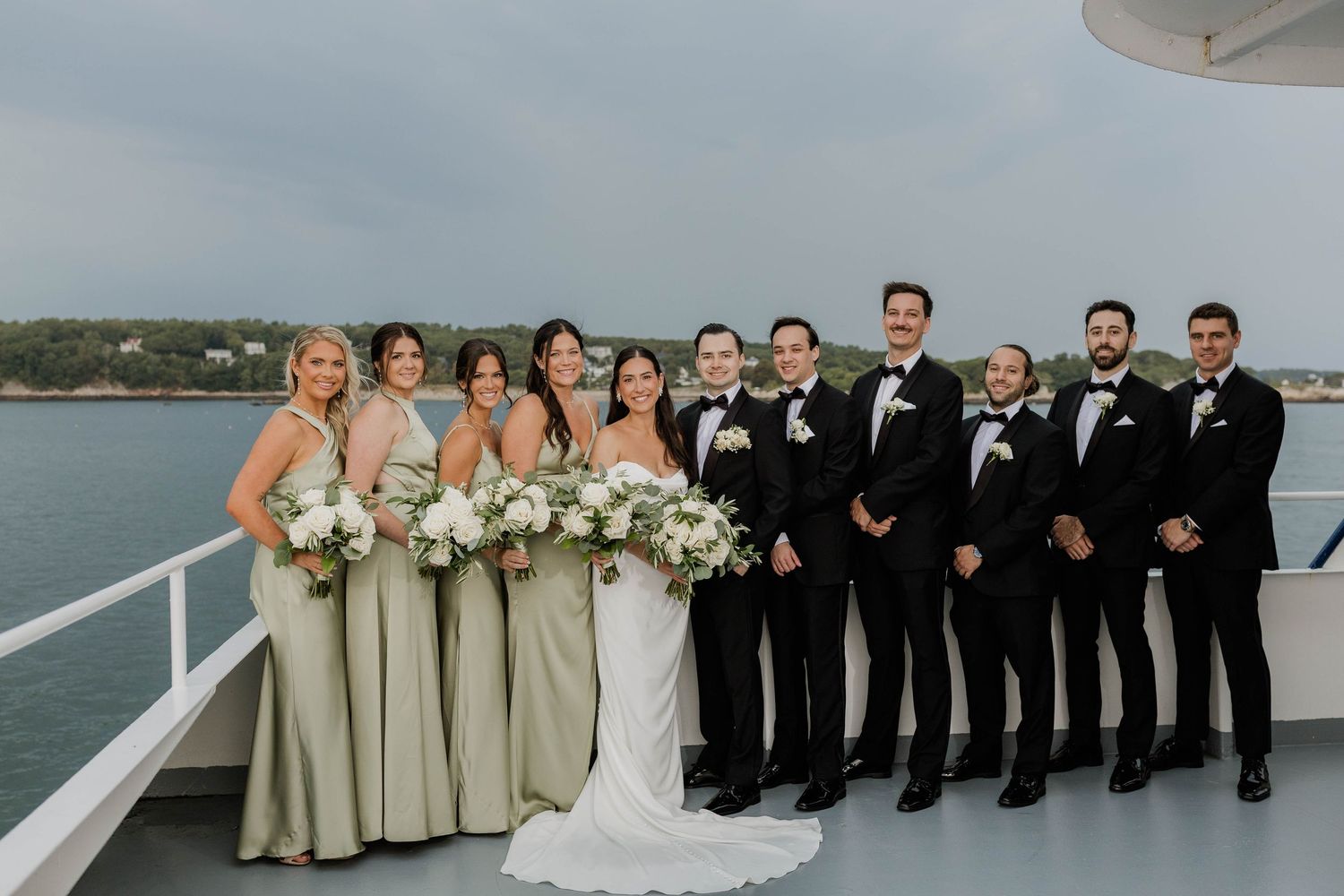 Wedding party poses on boat deck with bridesmaids in sage green and groomsmen in black tuxedos.