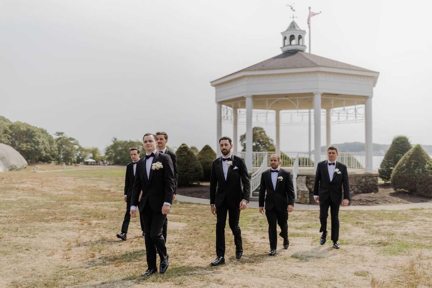 Groom and groomsmen in black tuxedos walking near white gazebo outdoors.