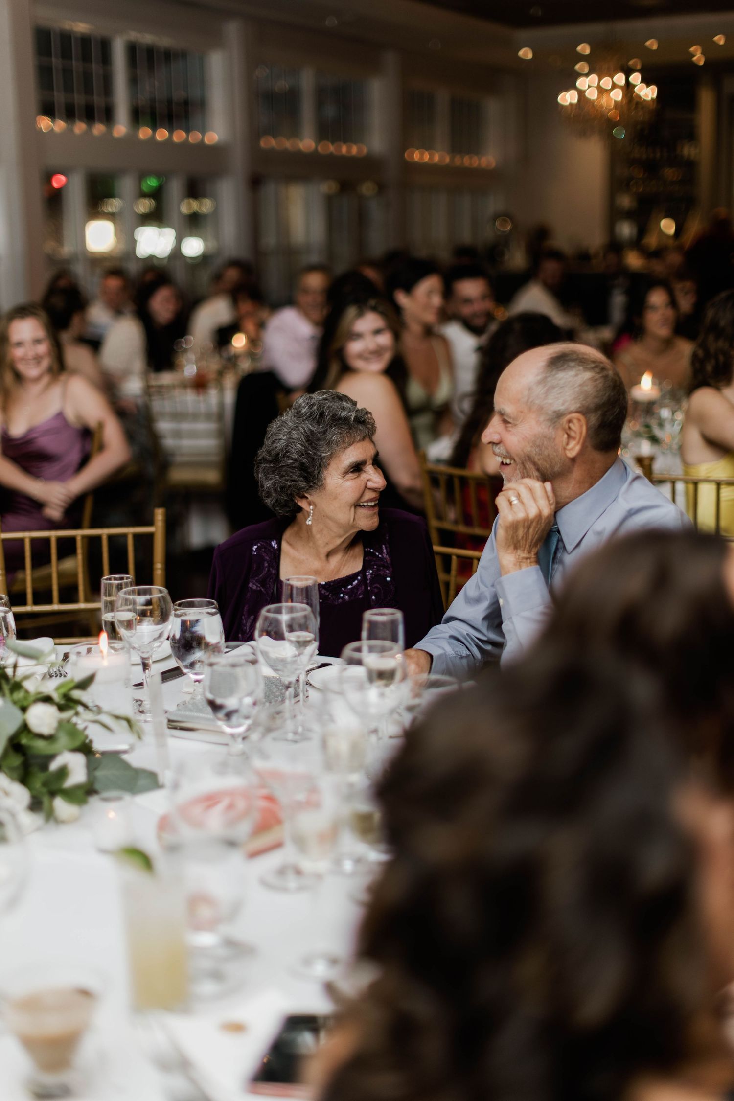 Guests laughing and smiling at wedding reception dinner table with candles and floral centerpieces.