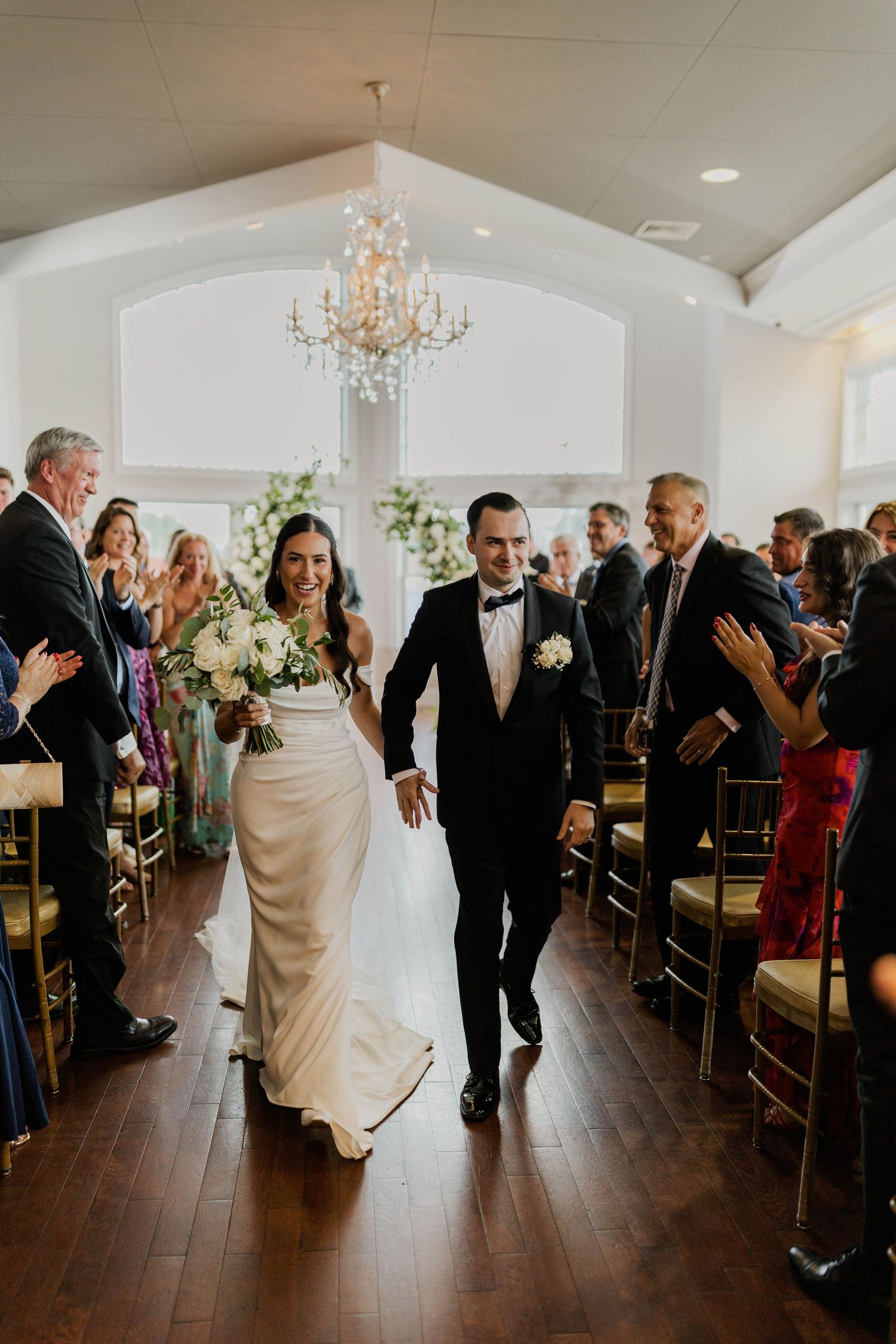 Newlyweds walk down aisle as guests applaud in bright white chapel with chandelier.