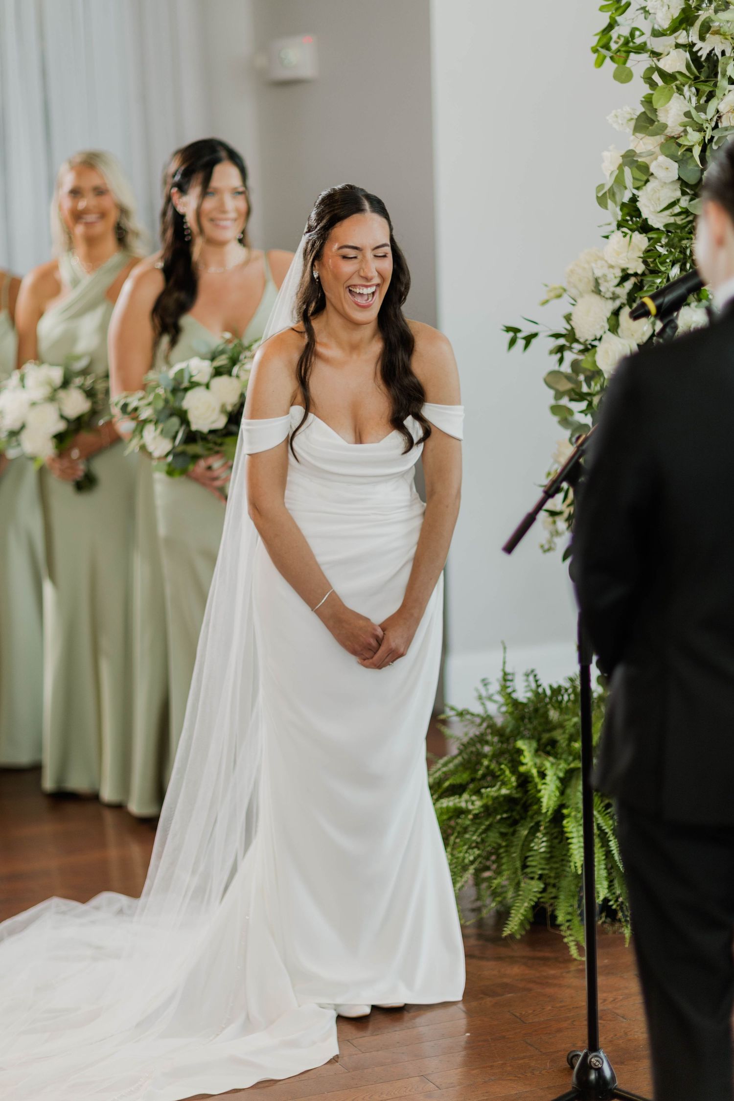 Radiant bride in an off-shoulder white gown laughing at the altar with bridesmaids in sage green.