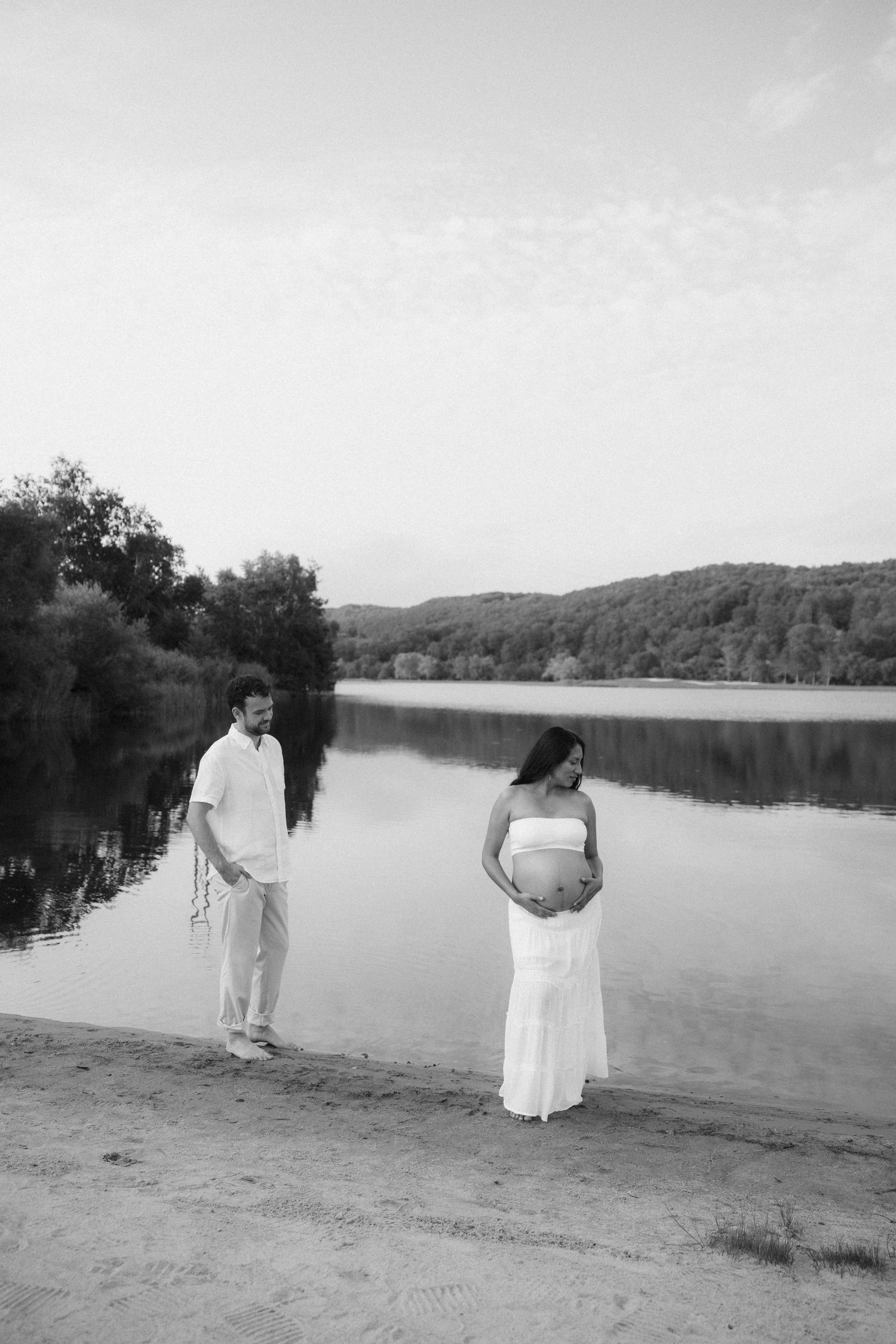 Black and white photo of a pregnant couple standing by a calm lake surrounded by trees.