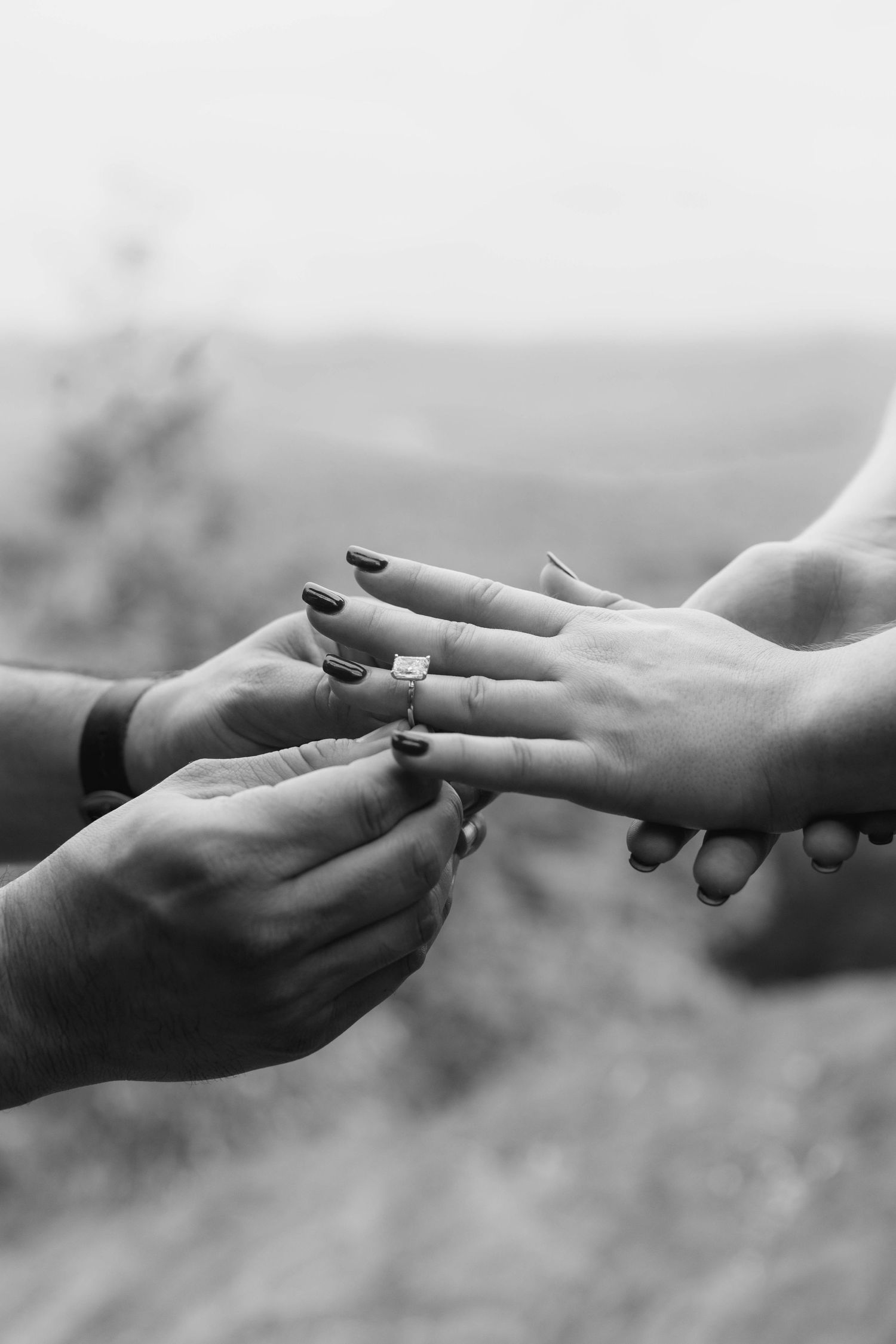 Black and white close-up of man sliding diamond engagement ring onto womans finger during proposal.