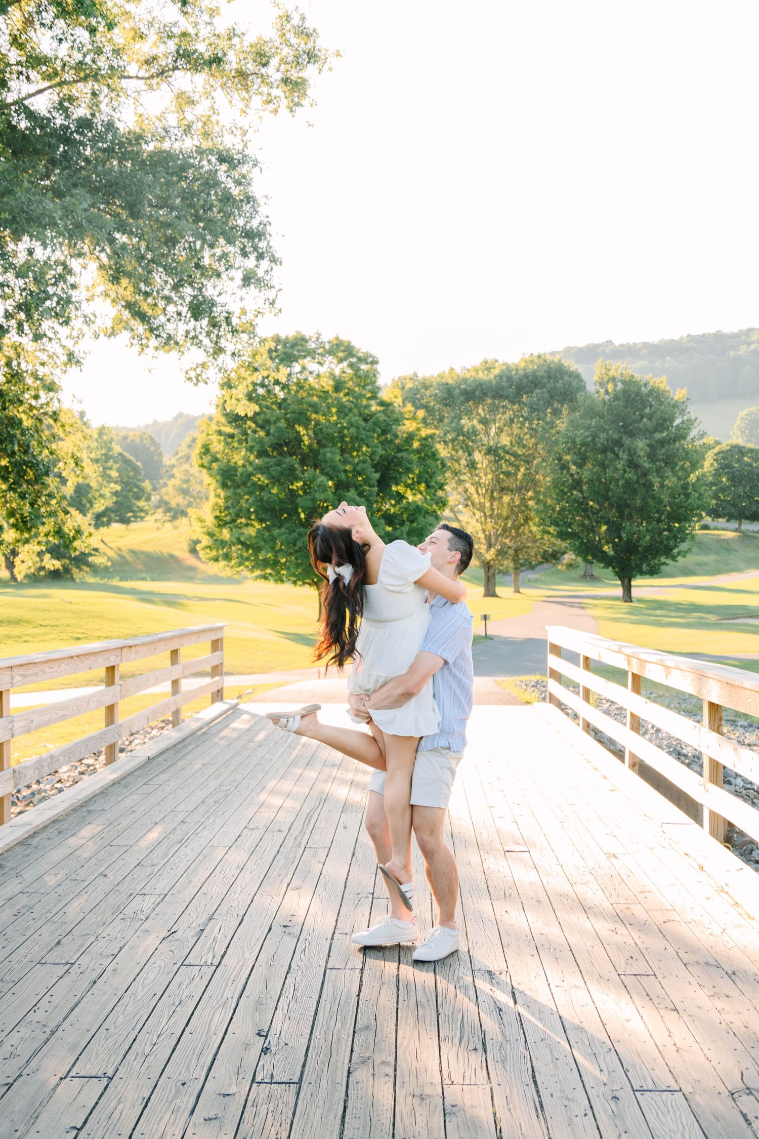 Couple sharing joyful moment on wooden bridge, man lifting woman in white dress during golden hour outdoors.
