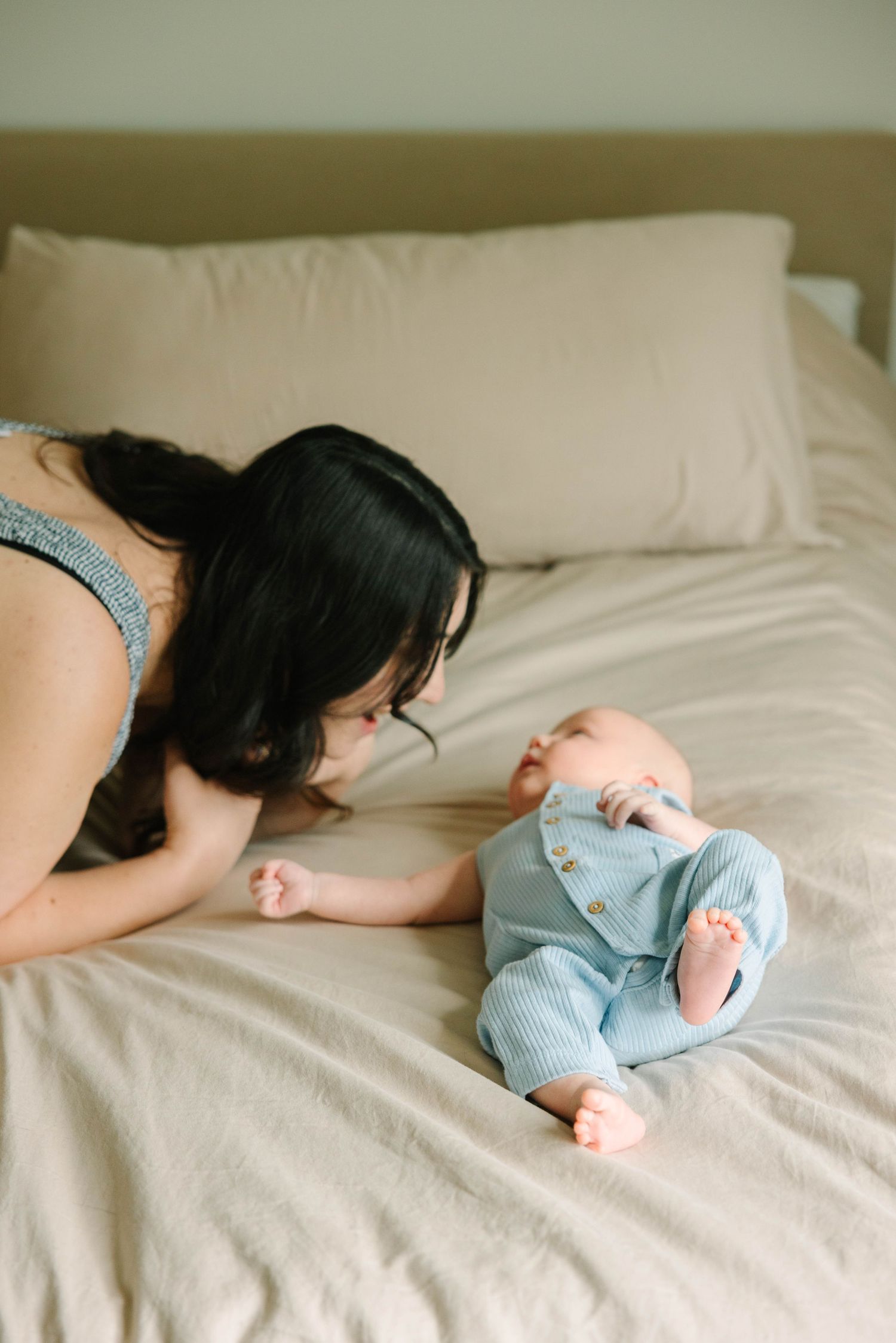 Young dark-haired mother leaning over her newborn baby in blue romper on a beige bed.