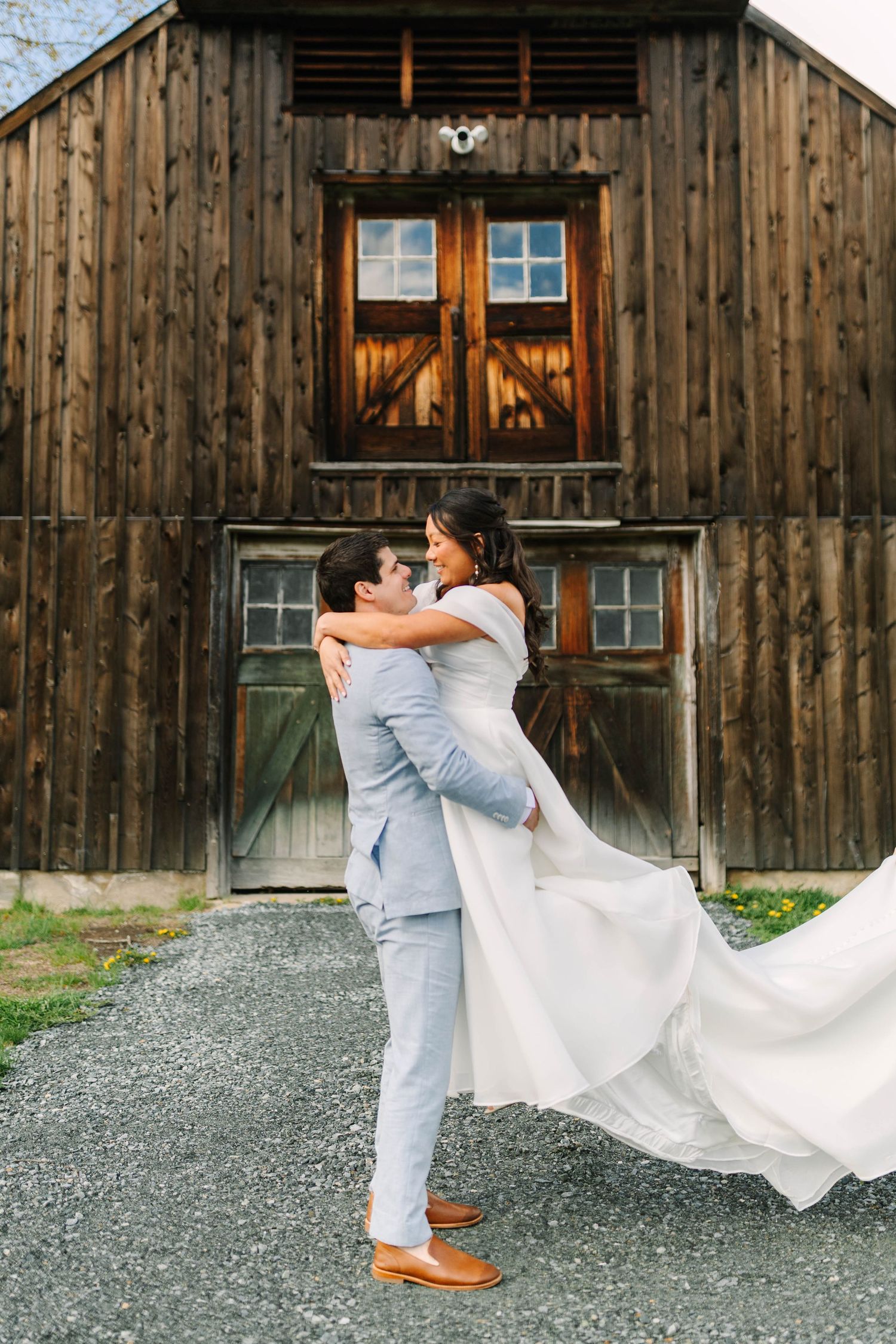 Groom in light blue suit lifts smiling bride in white gown outside rustic wooden barn.