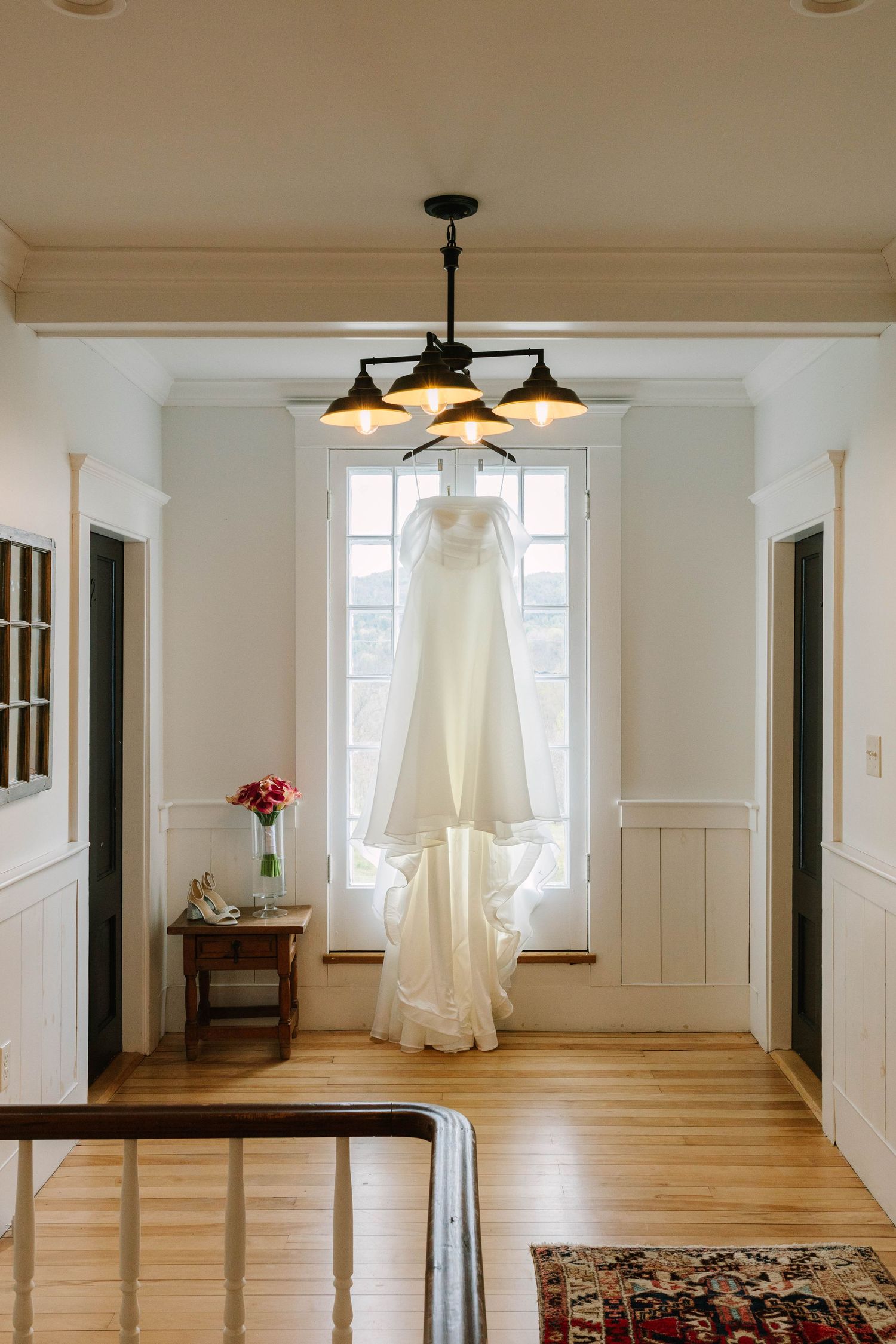 Elegant white wedding gown hanging from a chandelier in front of a bright window in a classic home.