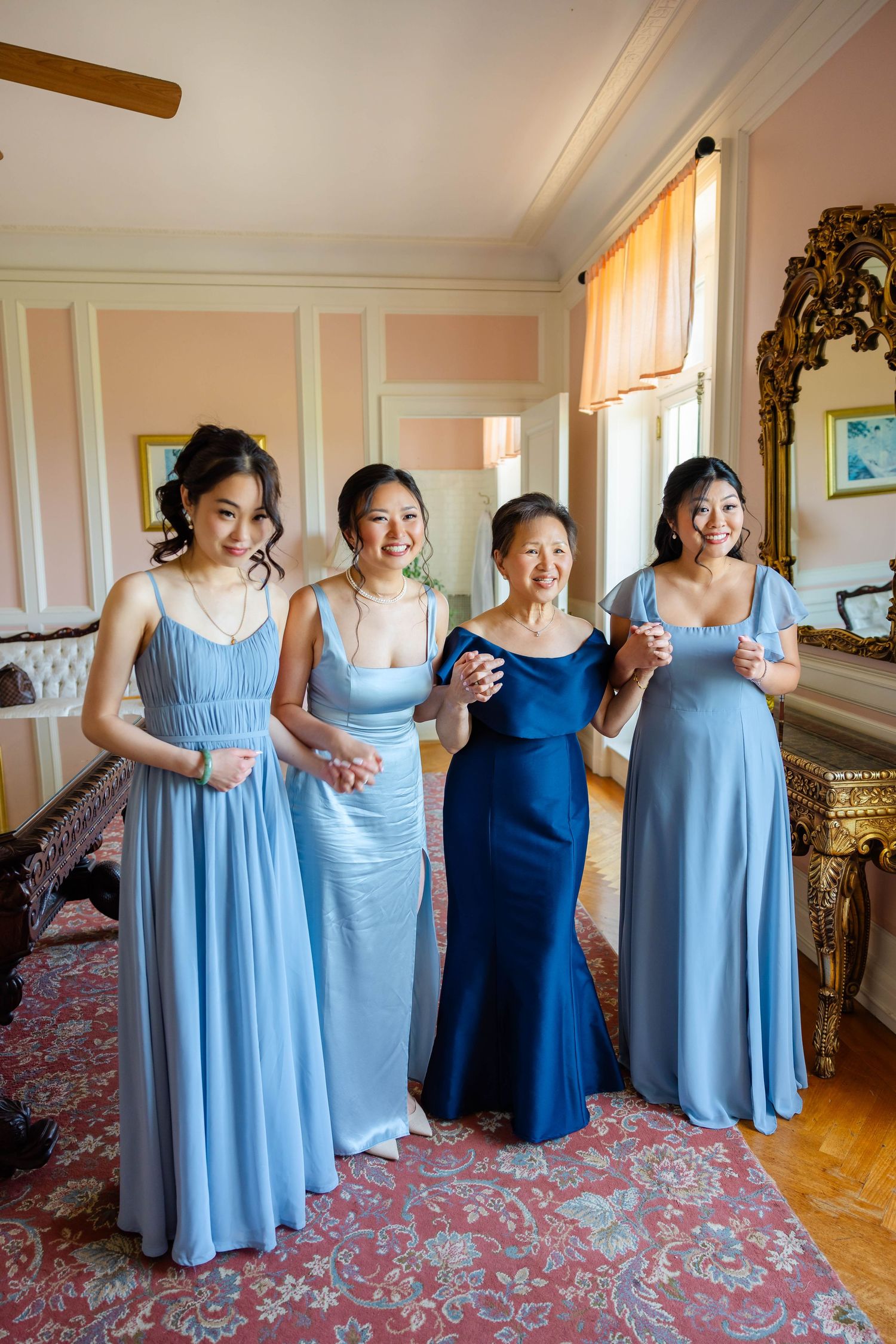 Four women in blue gowns smile together in an elegant pink room during wedding preparations.