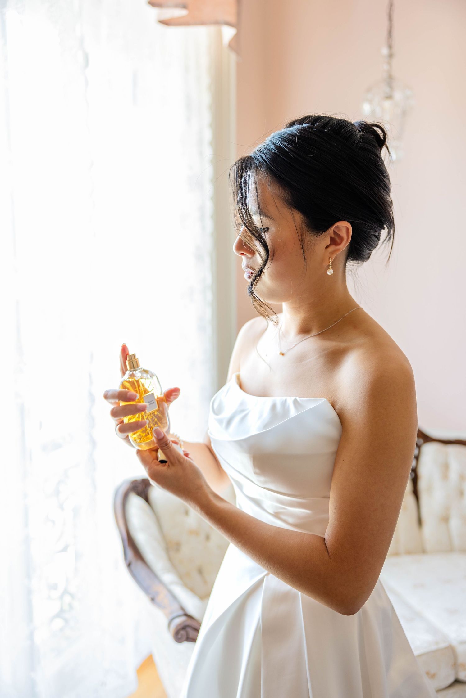 Bride in white strapless gown holding amber perfume bottle near bright window on wedding day.