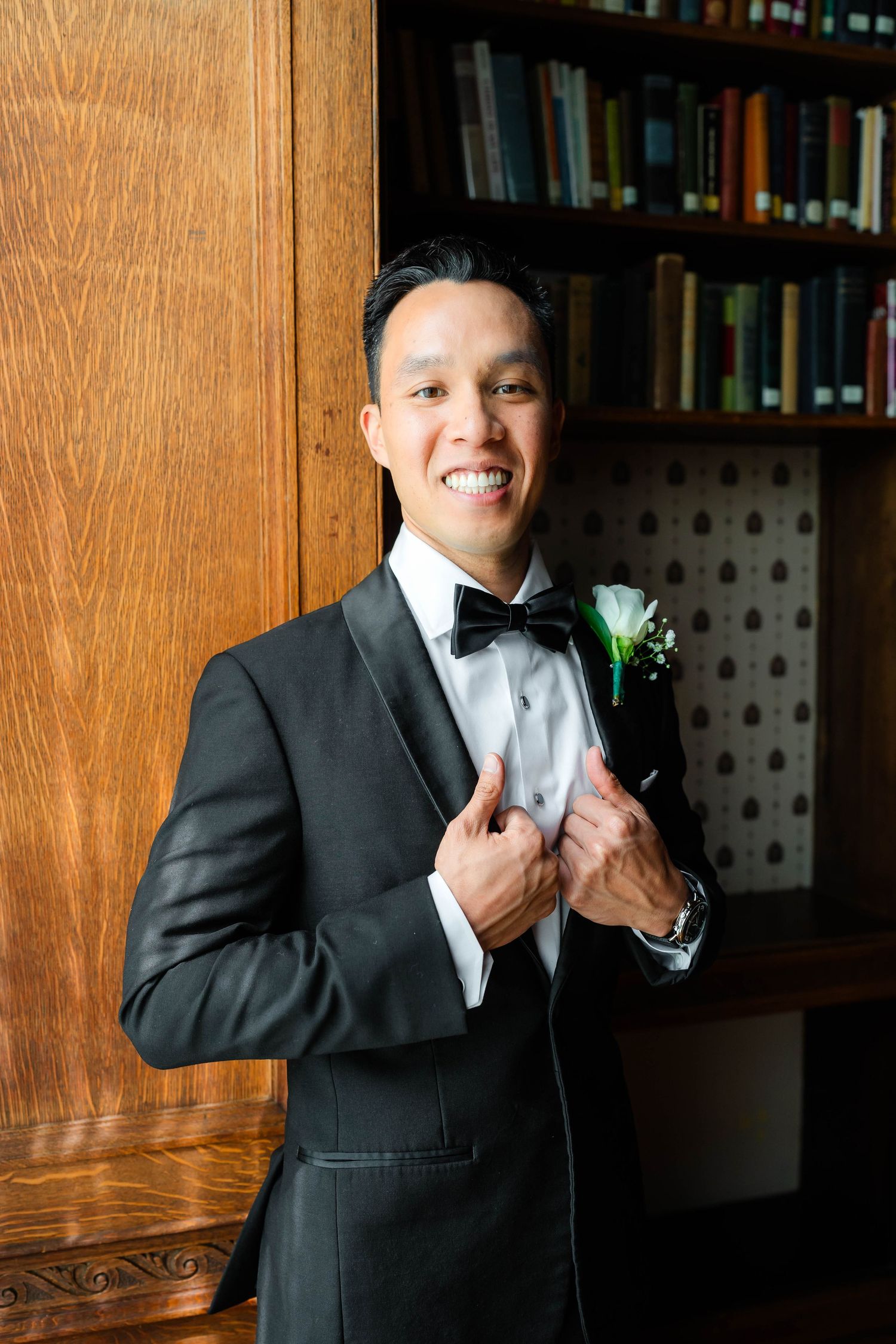 Smiling groom in black tuxedo with bow tie and boutonniere posing in a library.