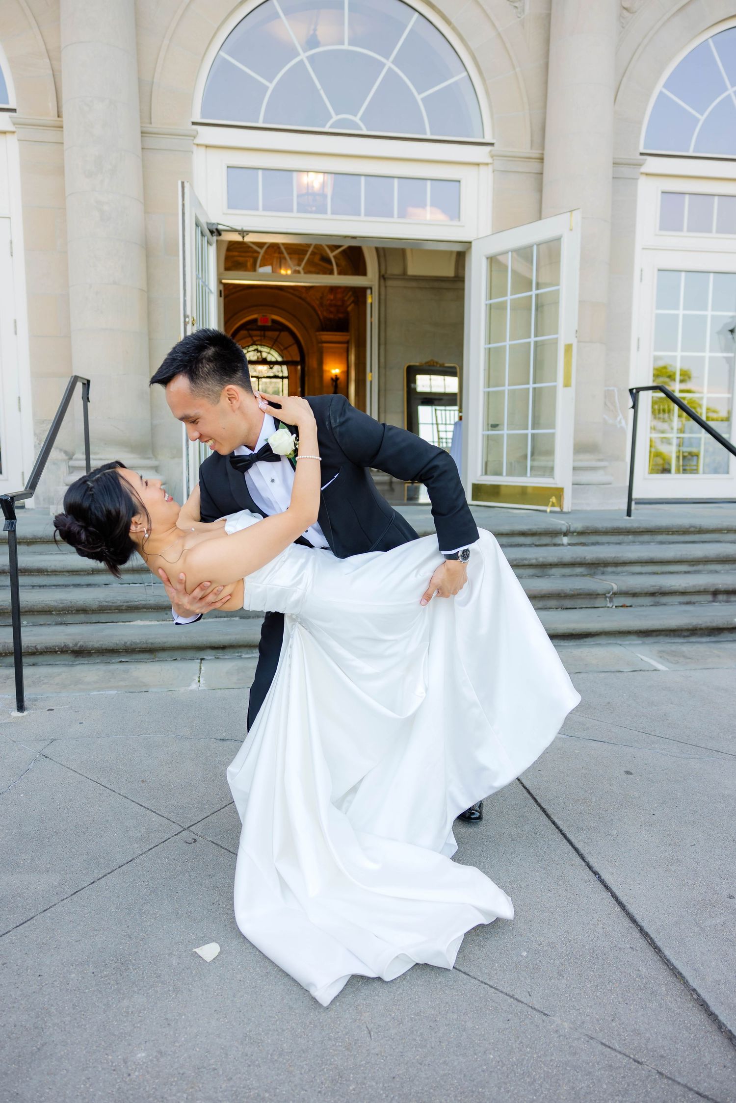 Groom dips bride in white wedding dress outside grand building entrance steps.