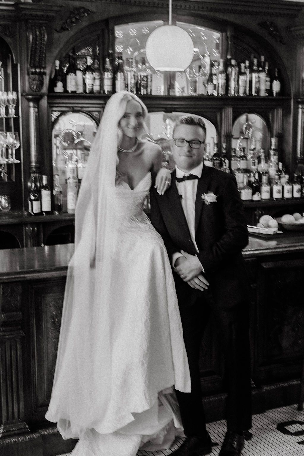 Black and white photo of bride and groom posing elegantly in front of a classic wooden bar.