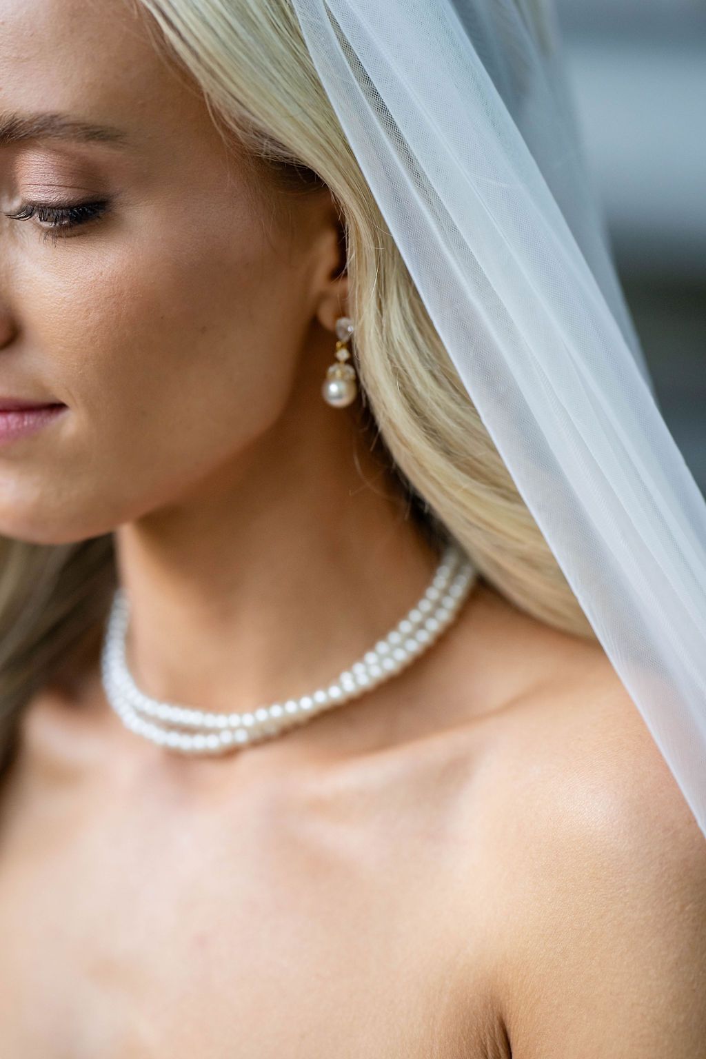 Close-up of bride wearing pearl necklace and earrings with a delicate white veil draped over blonde hair.