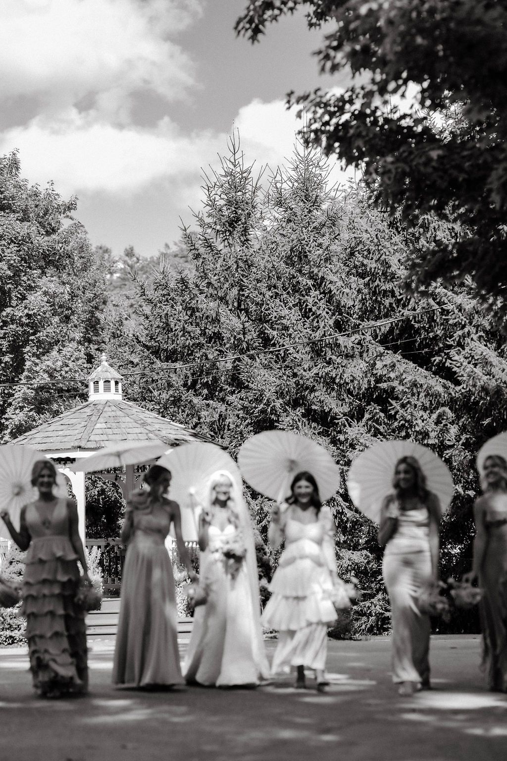 Black and white photo of a bride and bridesmaids holding parasols walking through a garden with a gazebo.