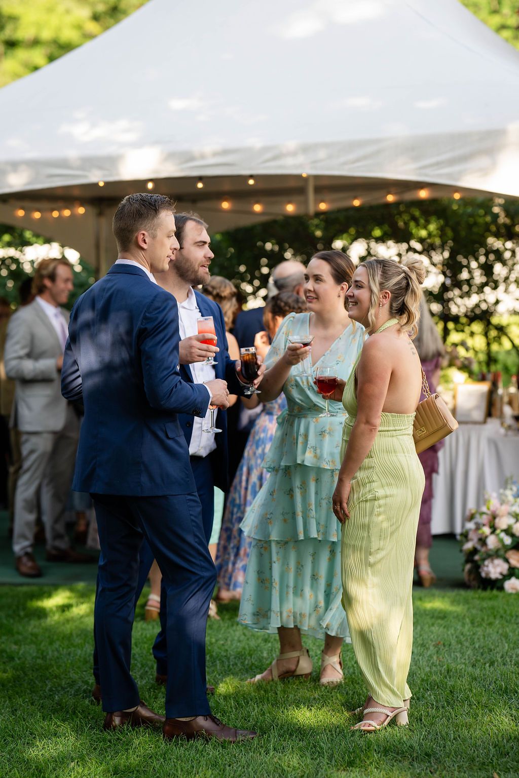 Guests in elegant attire socializing outdoors at a wedding reception under a white tent with string lights.