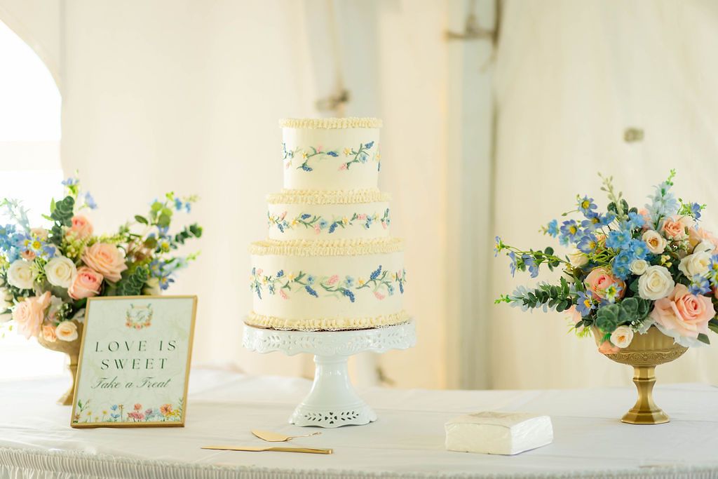 Three-tier white wedding cake with floral details displayed between pink and blue flower arrangements.