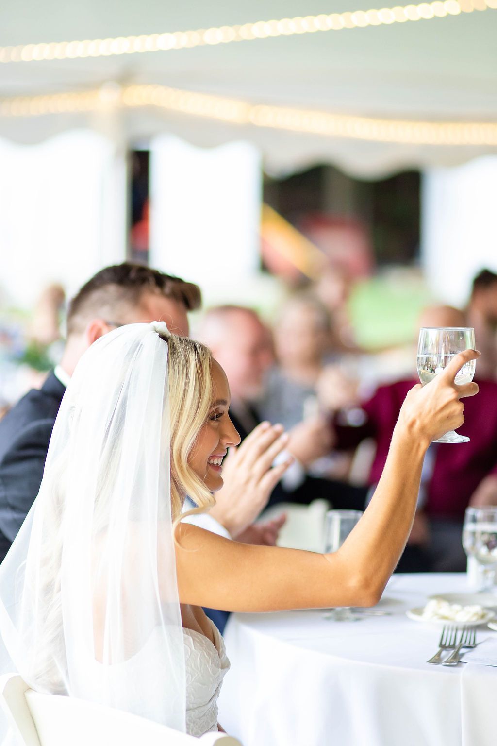 Smiling bride in white veil raises a glass of water during wedding reception toasts under a tent.