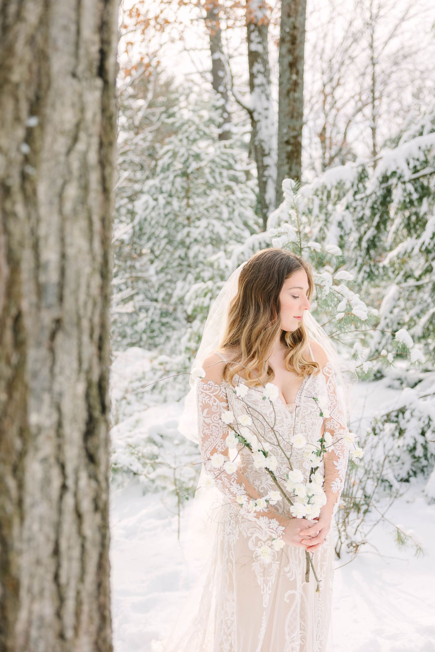 Bride in lace gown holding white floral bouquet in snowy winter forest.