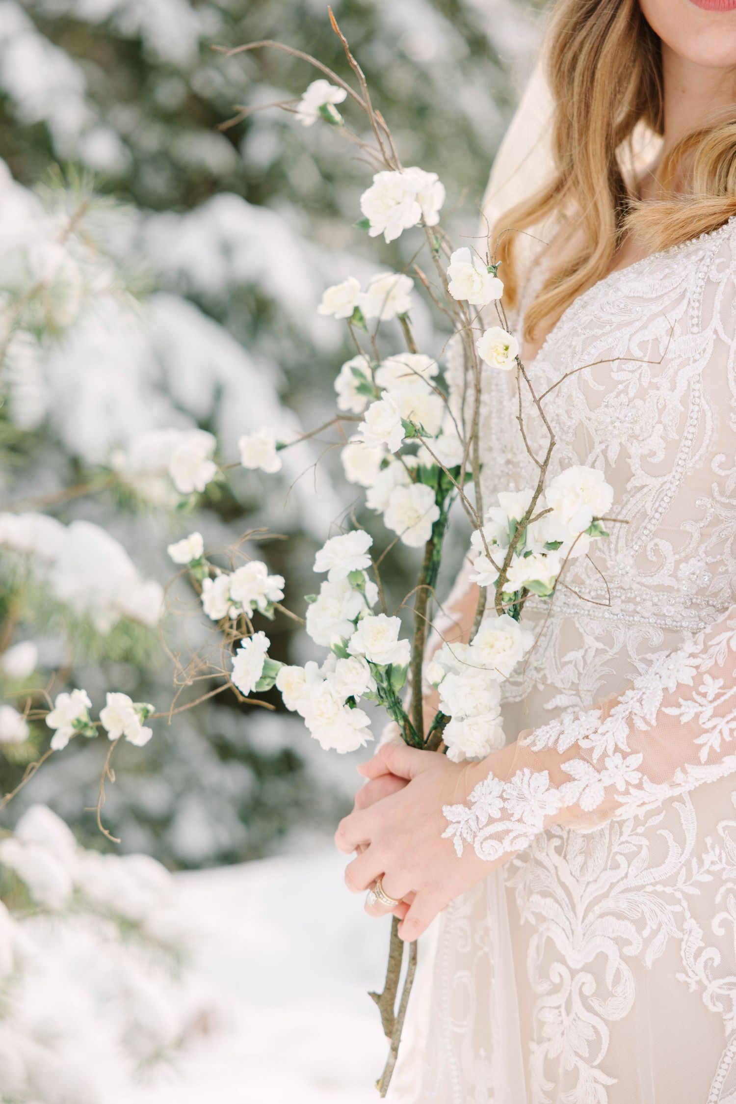 Bride in lace gown holding white floral branches in a snowy winter outdoor setting.