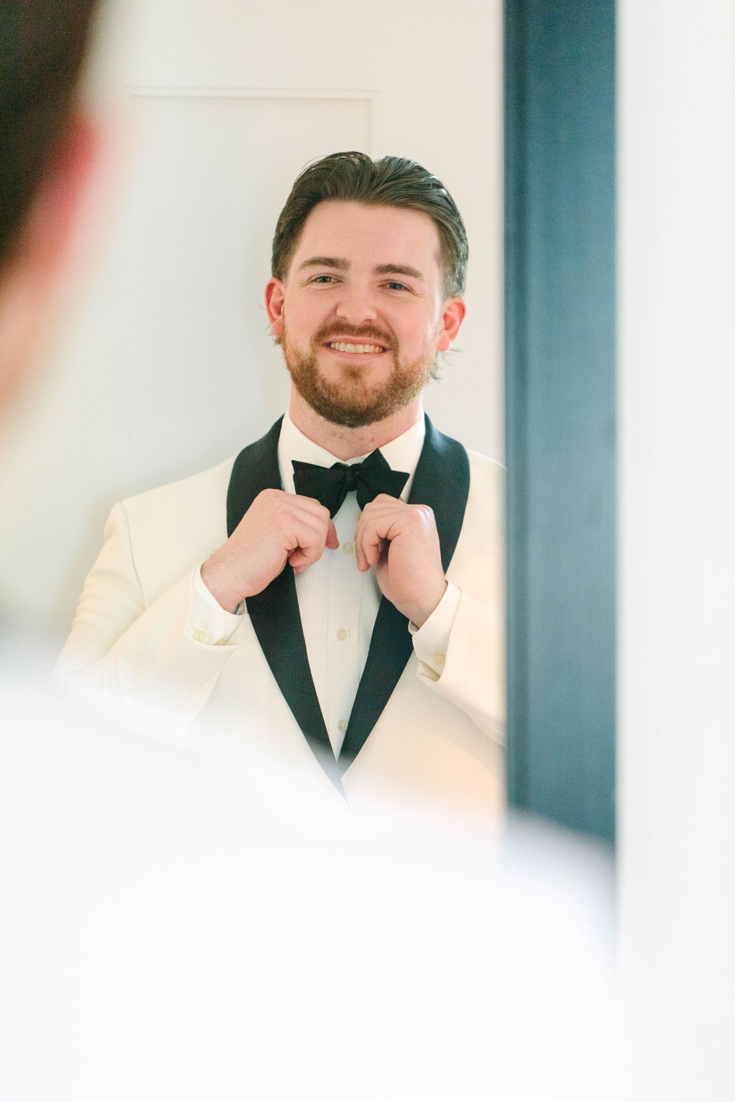 Handsome groom in ivory tuxedo adjusting his black bow tie while looking in a mirror.