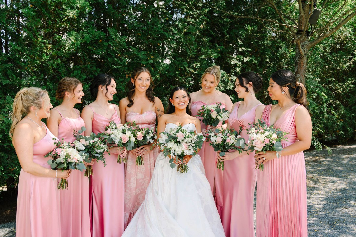 Smiling bride surrounded by bridesmaids in pink gowns holding pink and white floral bouquets.