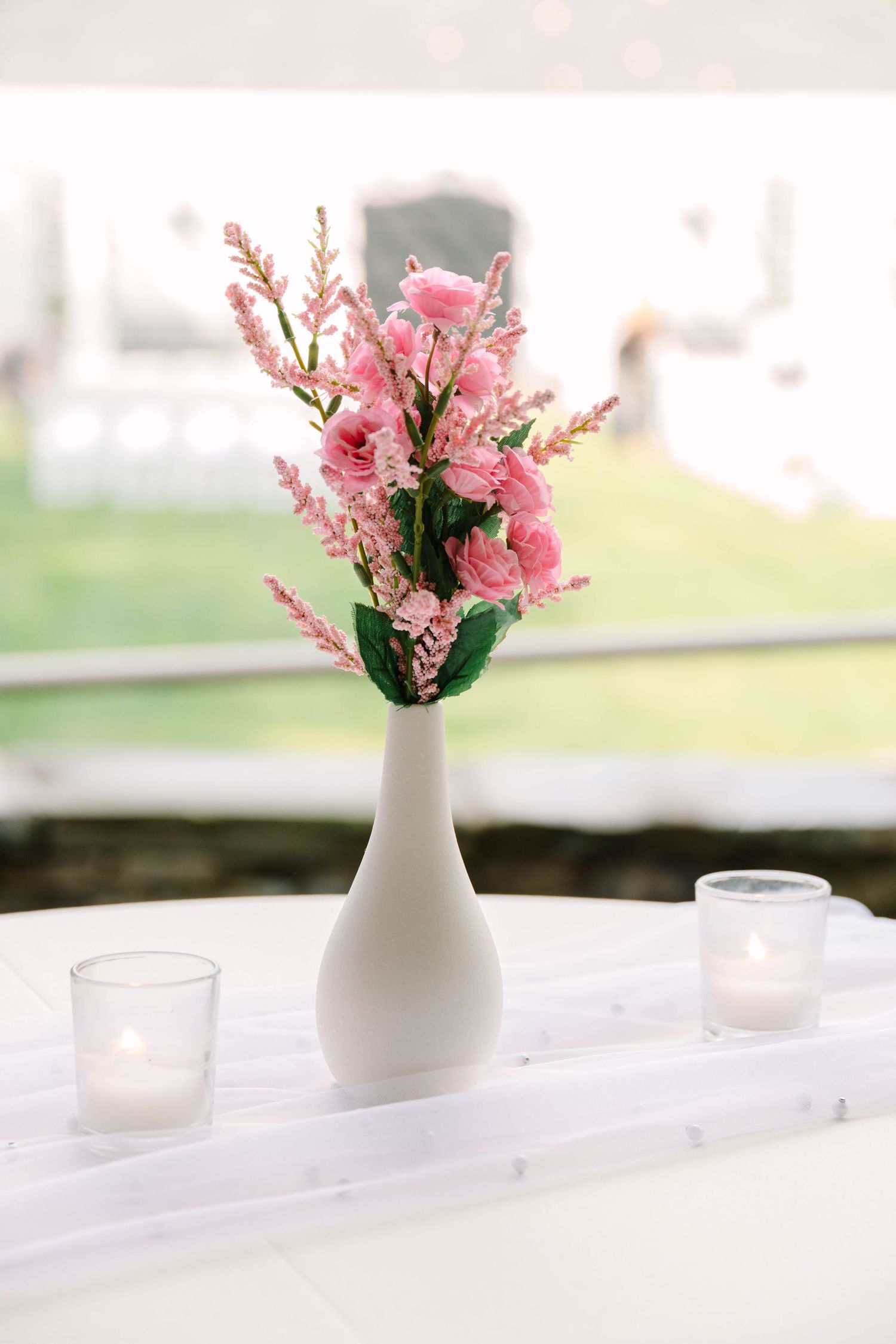 White bud vase with pink roses and snapdragons flanked by lit votive candles on white table runner.