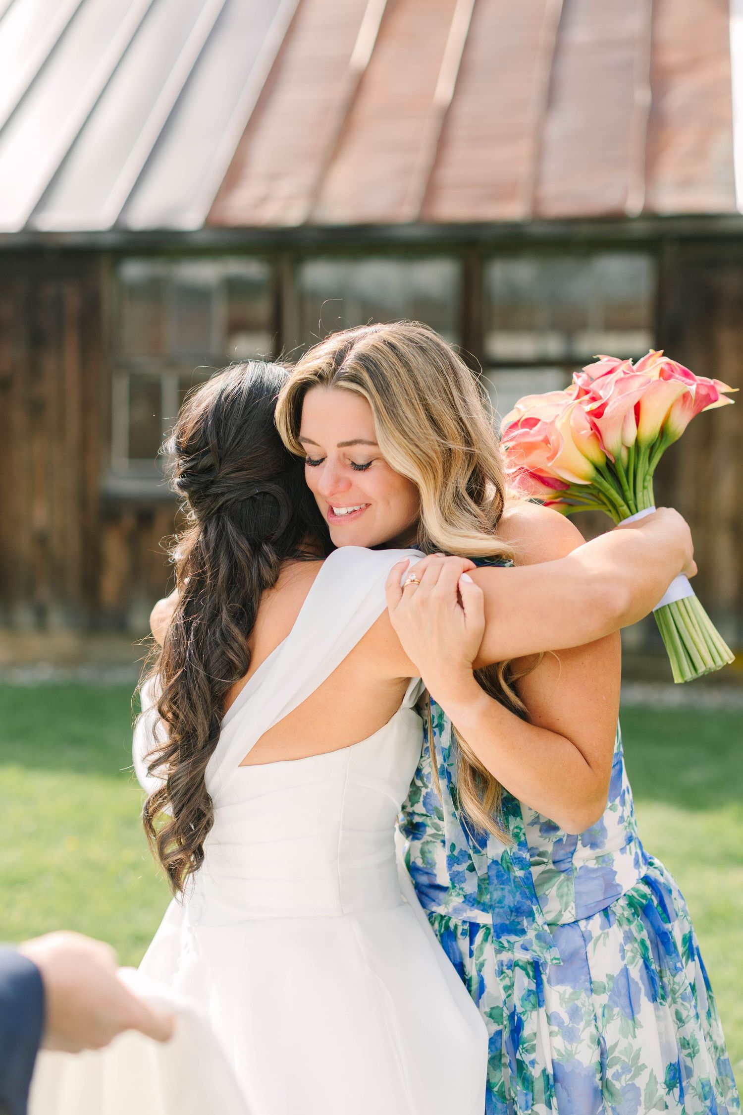 Bride in white backless gown embracing smiling blonde bridesmaid holding pink calla lily bouquet at outdoor barn wedding.