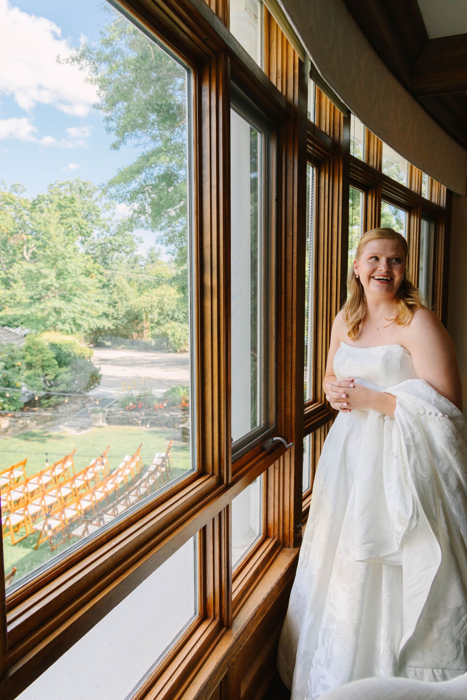 Radiant bride in a strapless white gown laughs by large wooden-framed windows overlooking outdoor ceremony chairs.