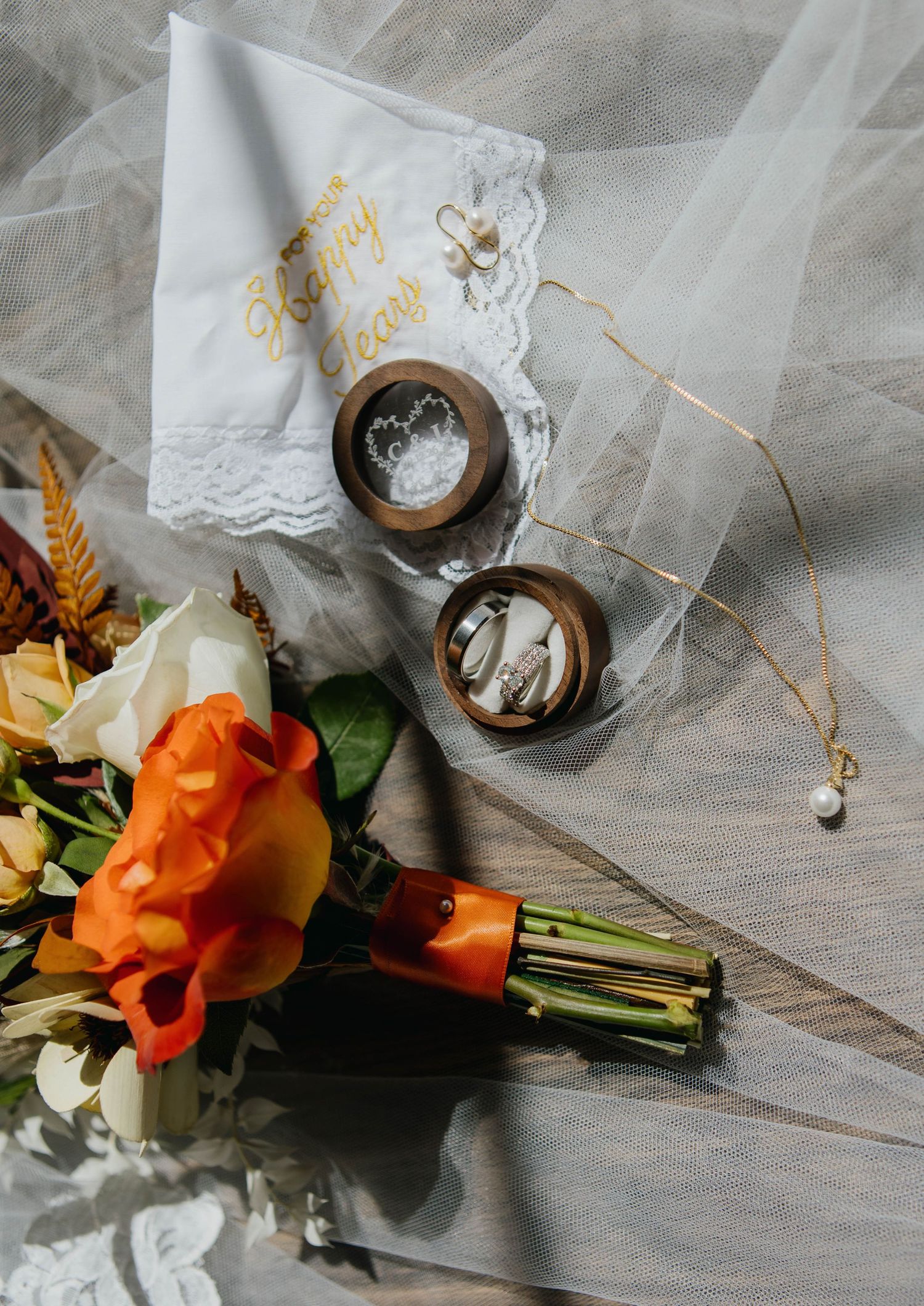Wedding flat lay with orange floral bouquet, gold rings in wooden boxes, lace handkerchief, and pearl necklace on veil.